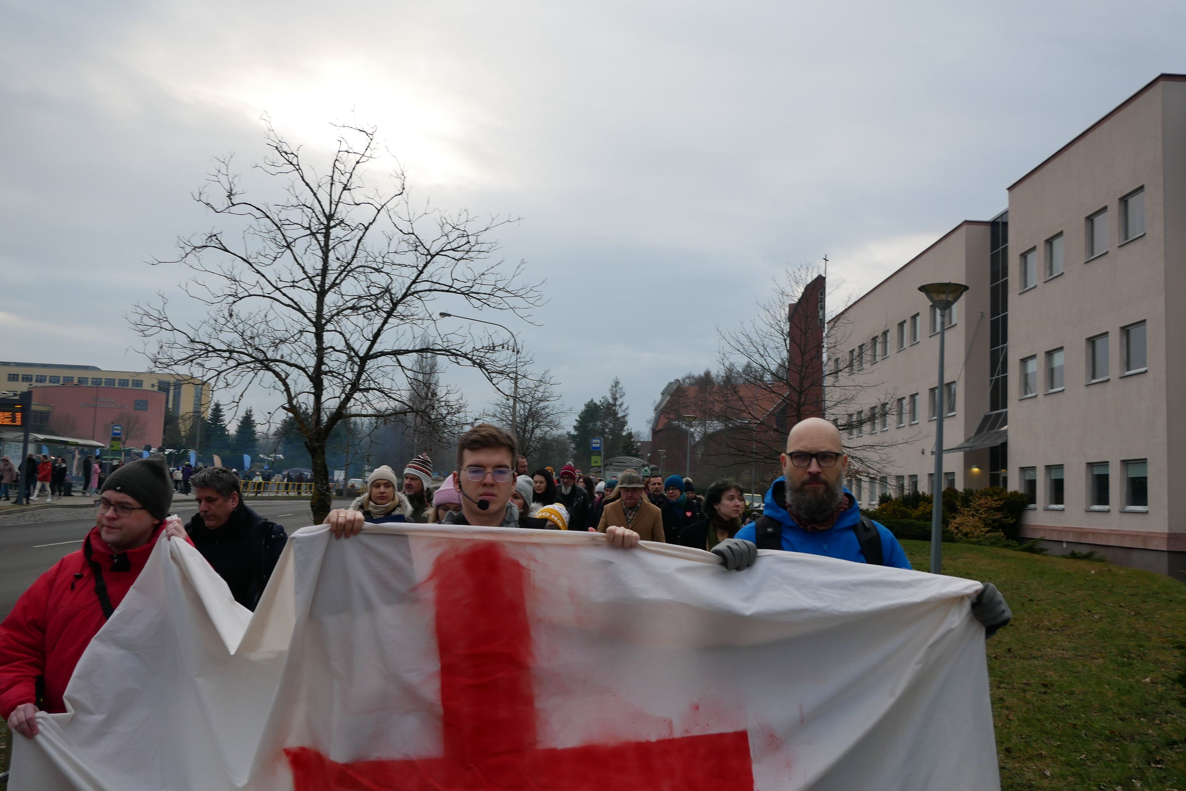 Protest uliczny, na pierwszym planie trzej młodzi ludzie niosą białe płótno z czerwonym krzyżem.