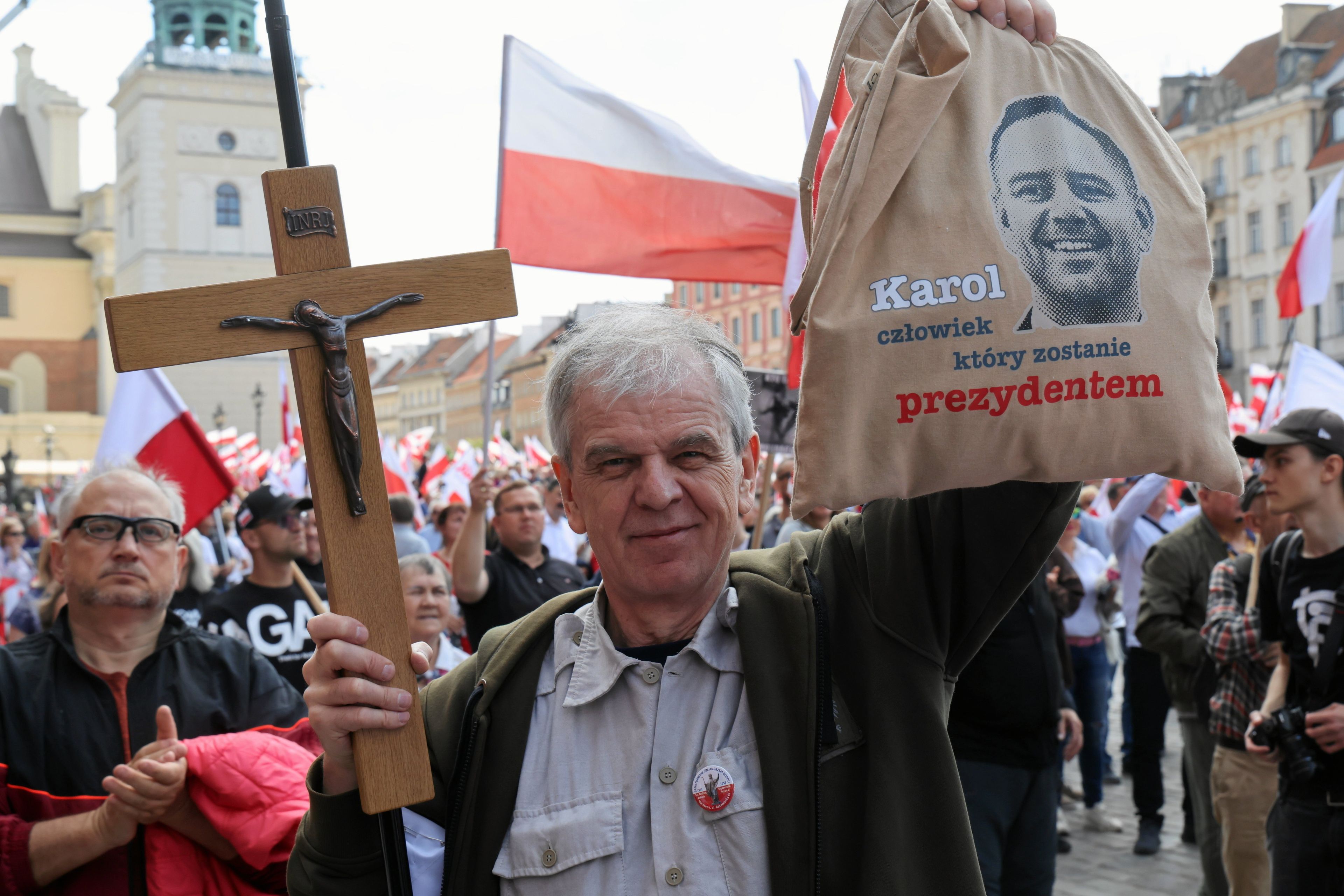 W tłumie ludzi z biało-czerwonymi flagami stoi starszy mężczyzna, w jednej ręce trzyma krzyż, a w drugiej torbę z podobizną Karola Nawrockiego i napisem "Karol, człowiek, który zostanie prezydentem"