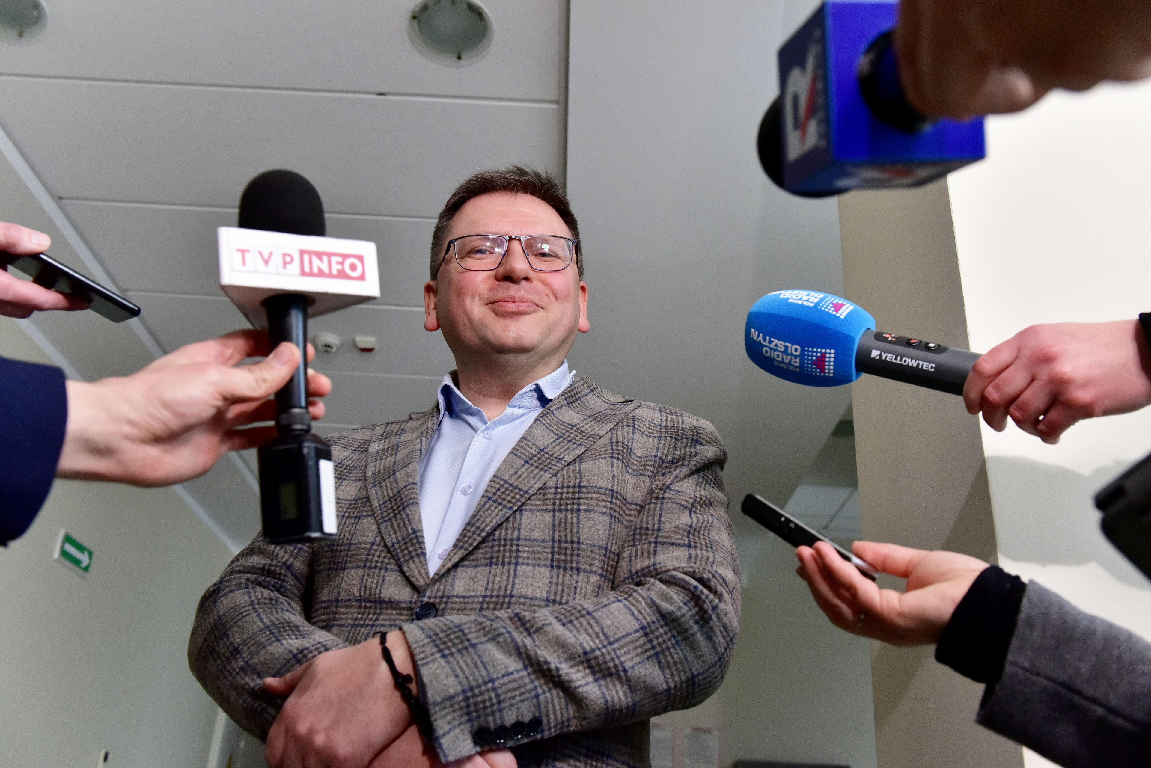 Mężczyzna w okulrach i kraciastej marynarce stoi przed dziennikarzami z uśmieszkiem na twarzy