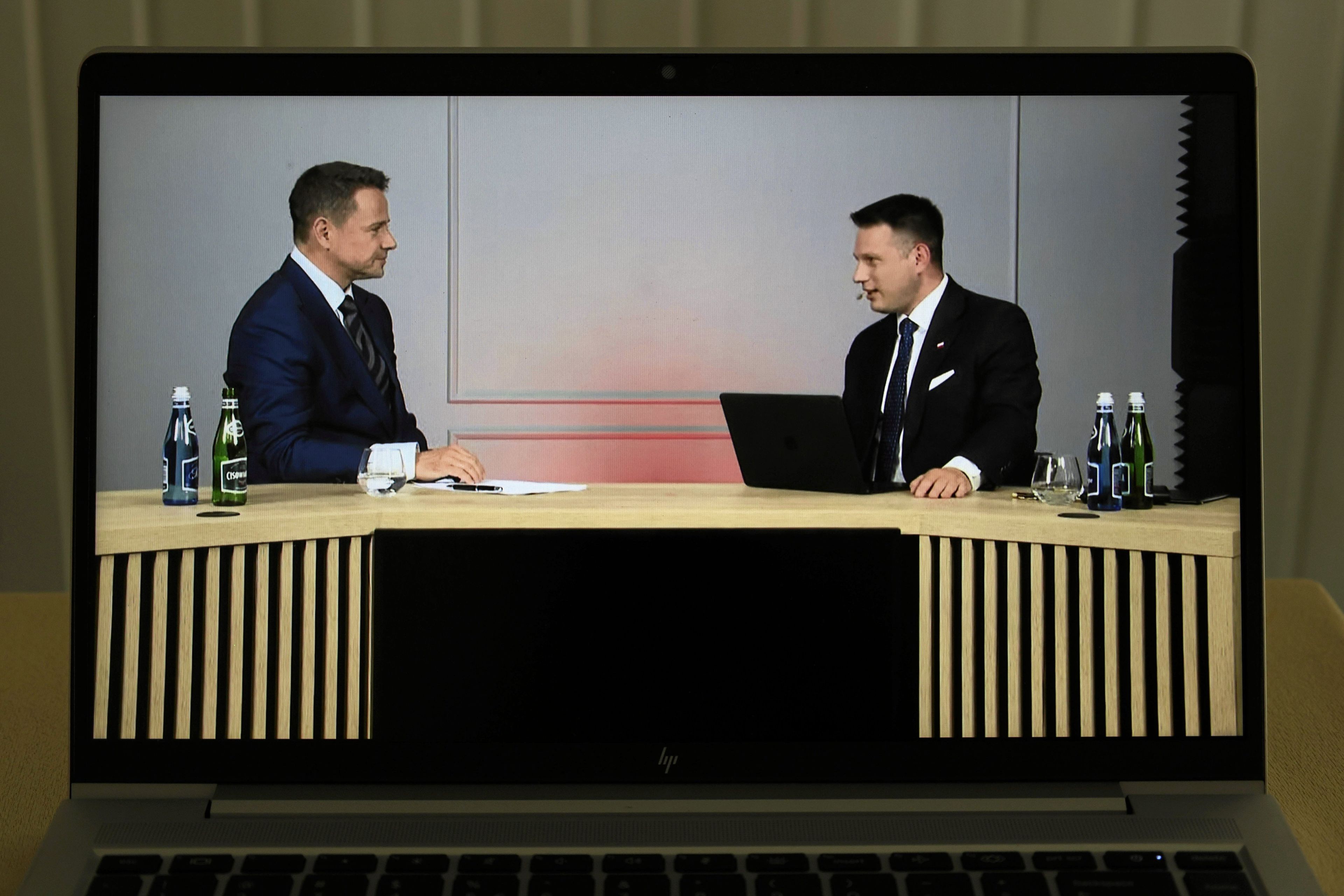 Dwóch mężczyzn w garniturach siedzi przy drewnianym stole w studiu nagraniowym, rozmawiają podczas wywiadu lub debaty. Każdy ma przed sobą butelki wody i szklankę. Jeden używa laptopa. Obraz widoczny jest na ekranie laptopa.
