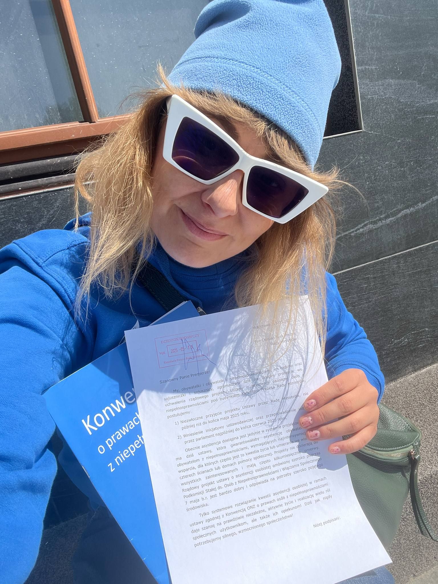 Kobieta ubrana na niebiesko, w dużych okularach, z książką w niebieskiej okładce i kartką zapisanego papieru