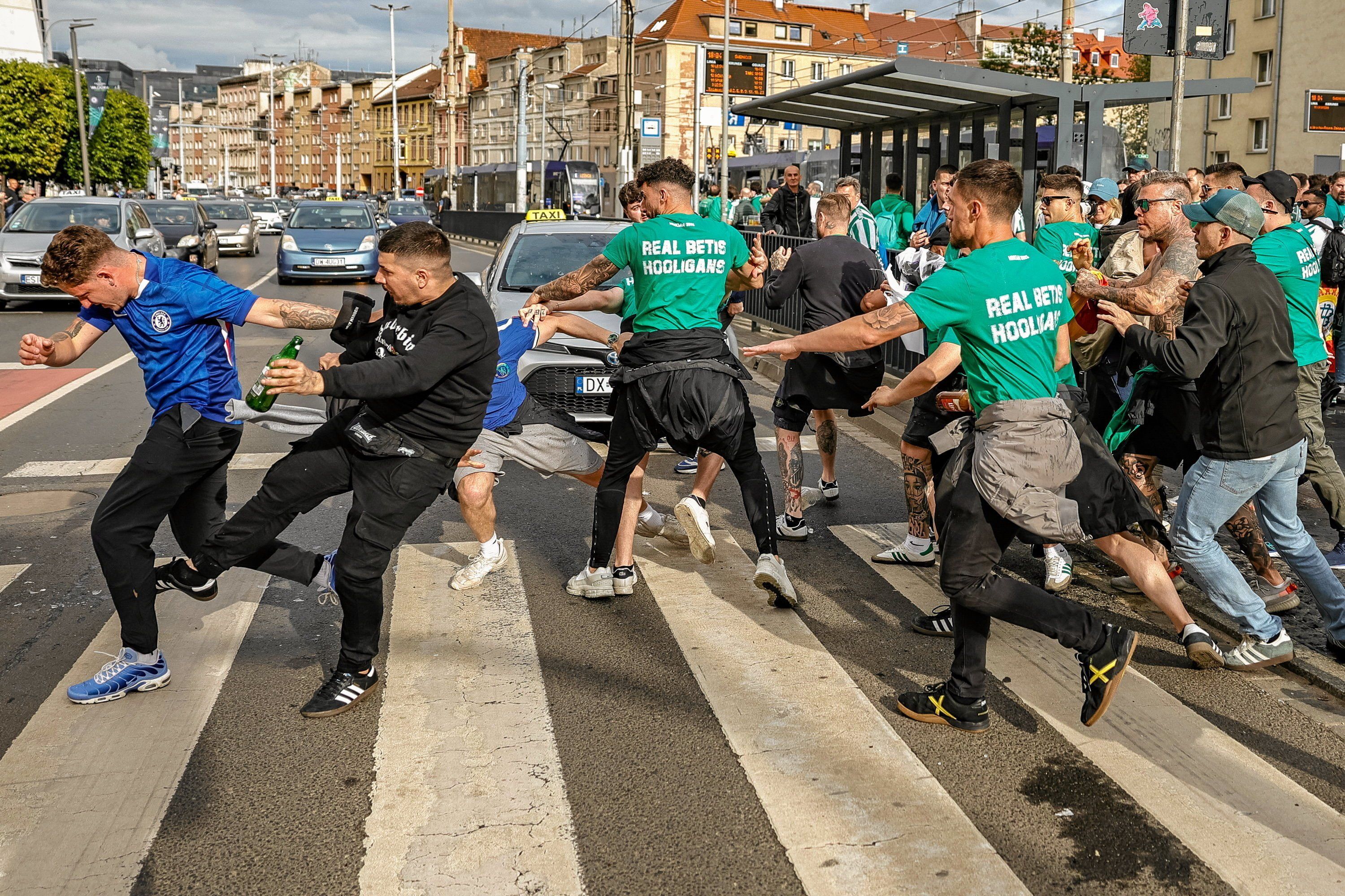 Grupa młodych mężczyzn w zielonych koszulkach z napisem "Real Betis Hools" na plecach szarpie i kopie grupę mężczyzn biegnących przez przejście dla pieszych
