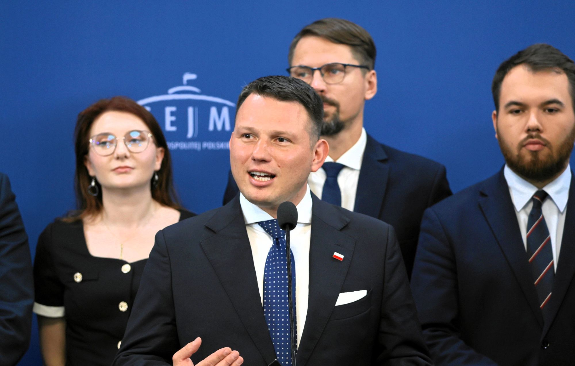 Mężczyzna w średnim wieku przemawia, za nim stoi kilka osób, w tle logo Sejmu.