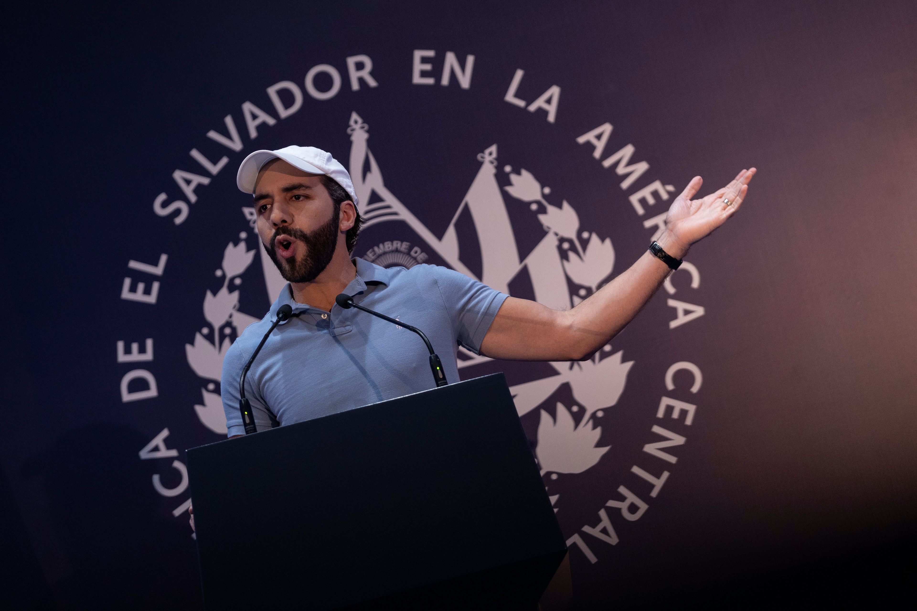 Młody mężczyzna w białej bejsbolówce i błękitnej koszulce przemawia z mównicy. Za nim napis: "Repuclica de el Salvador en la America Central