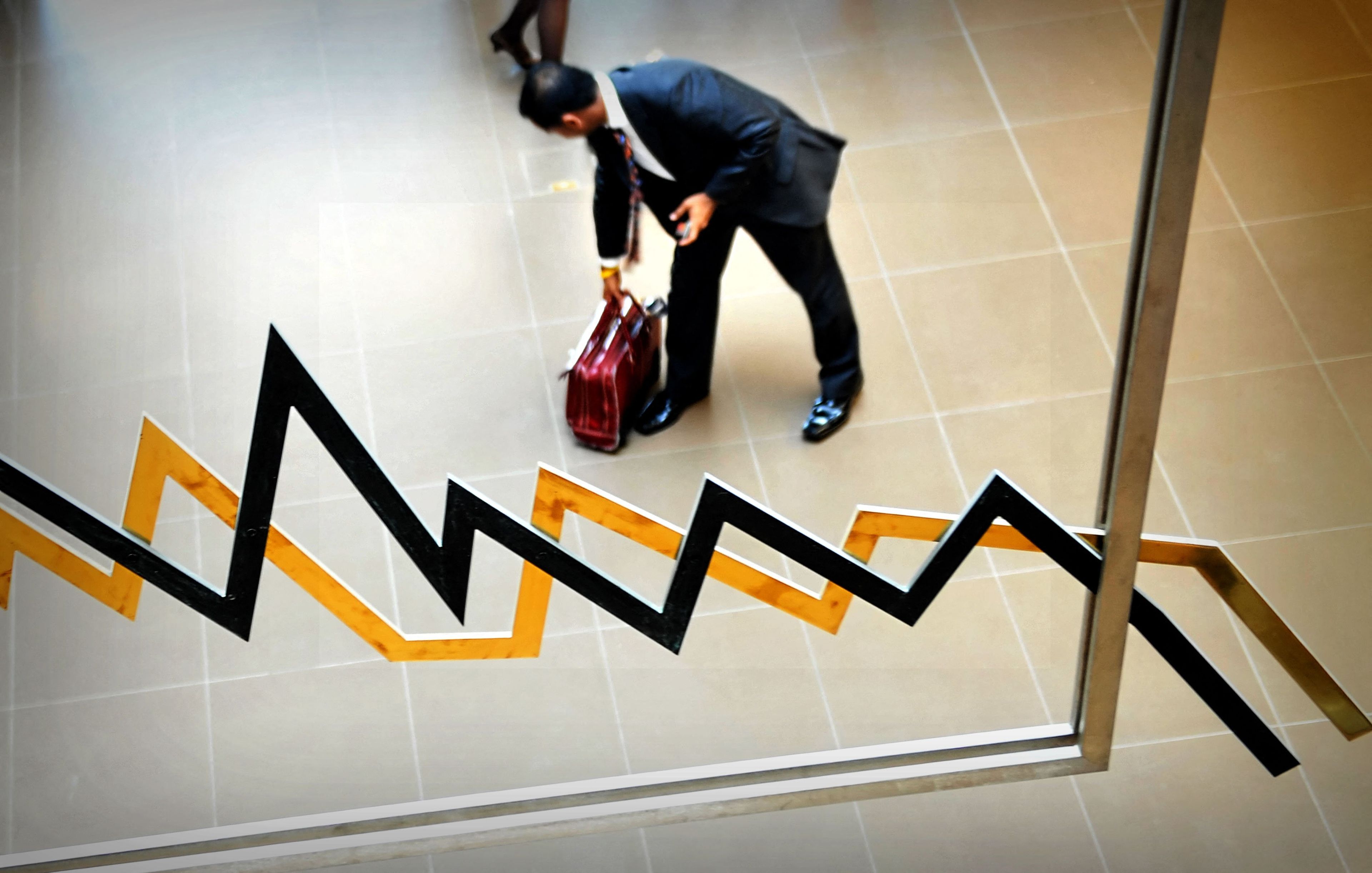 Mężczyzna podnosi torbę. Na pierwszym planie instalacja przypominająca wykres kursów giełdowych