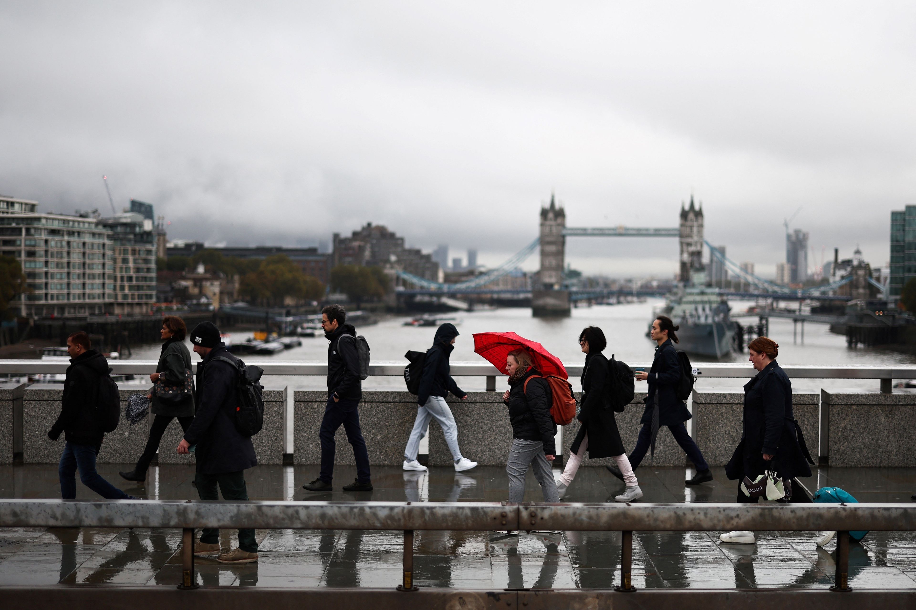 Piesi idą jeden za drugim po moście. W tle londyński Tower Bridge