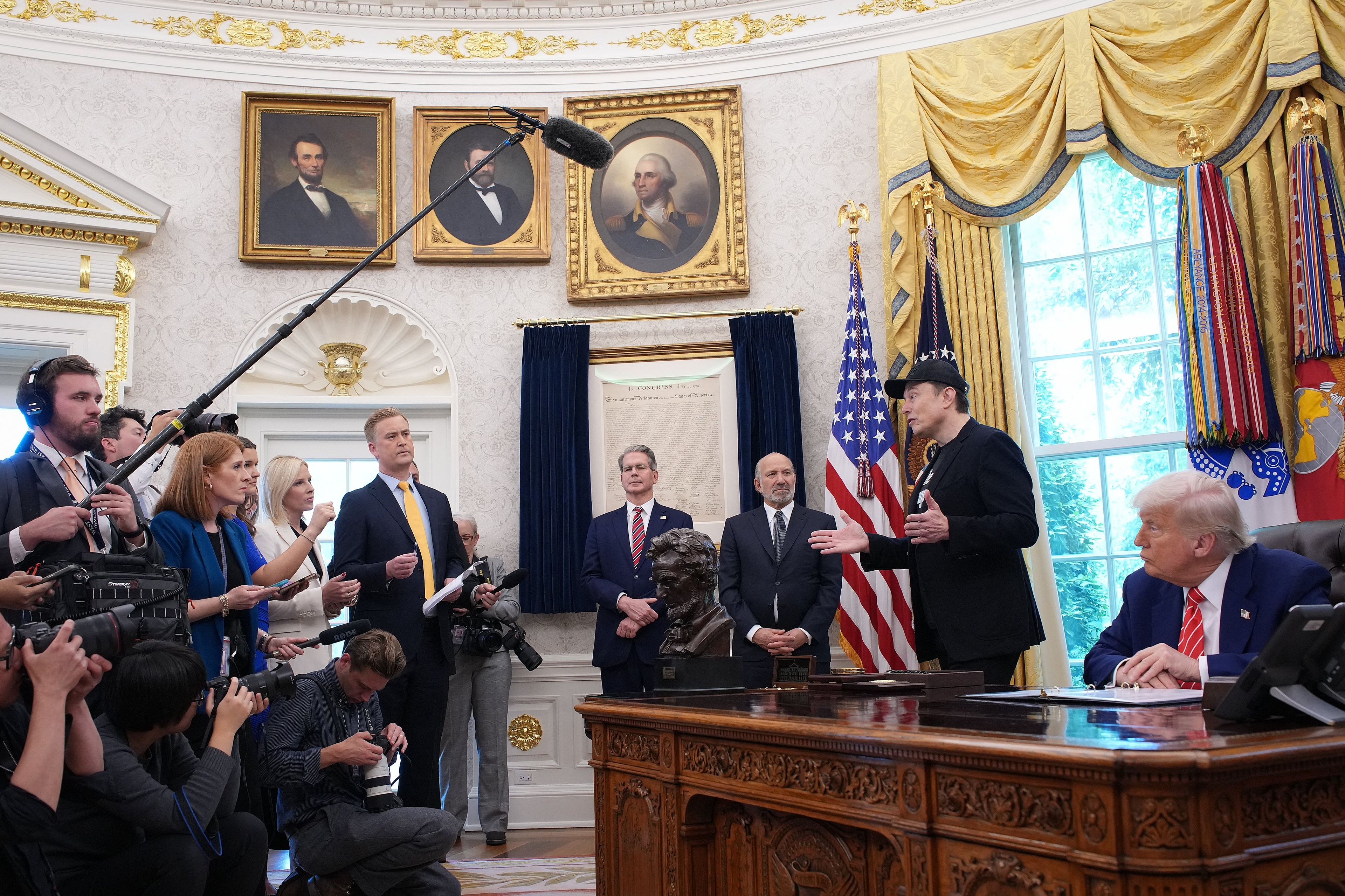 Mężczyzna w czapce z daszkiem (Musk) stoi obok Trumpoa siedzącego przy prezydenckim biurku w Gabinecie Owalnym. Naprzeciwko - dziennikarze
