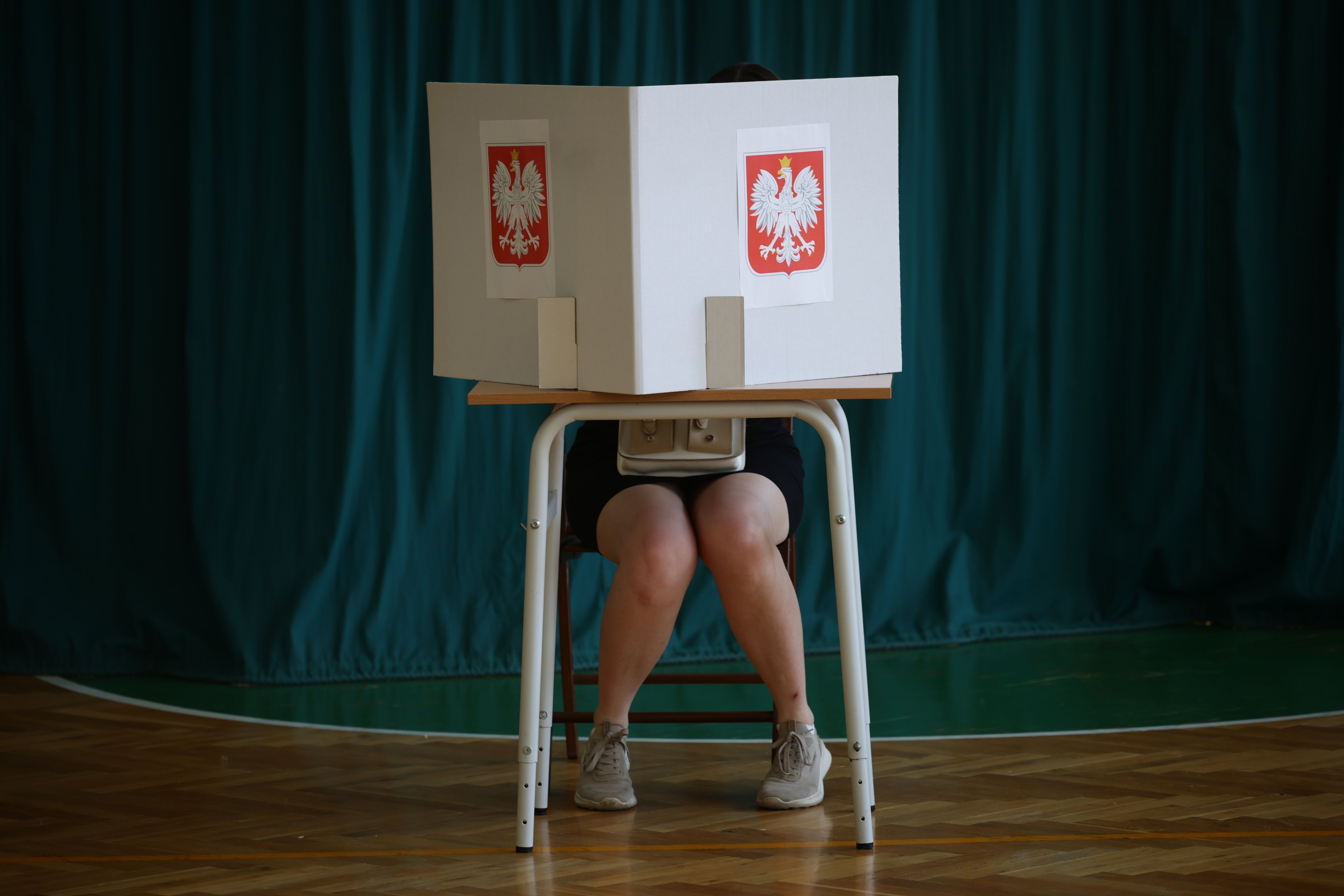 Osoba siedząca przy stoliku zasłoniętym przesłoną z polskim godłem.
