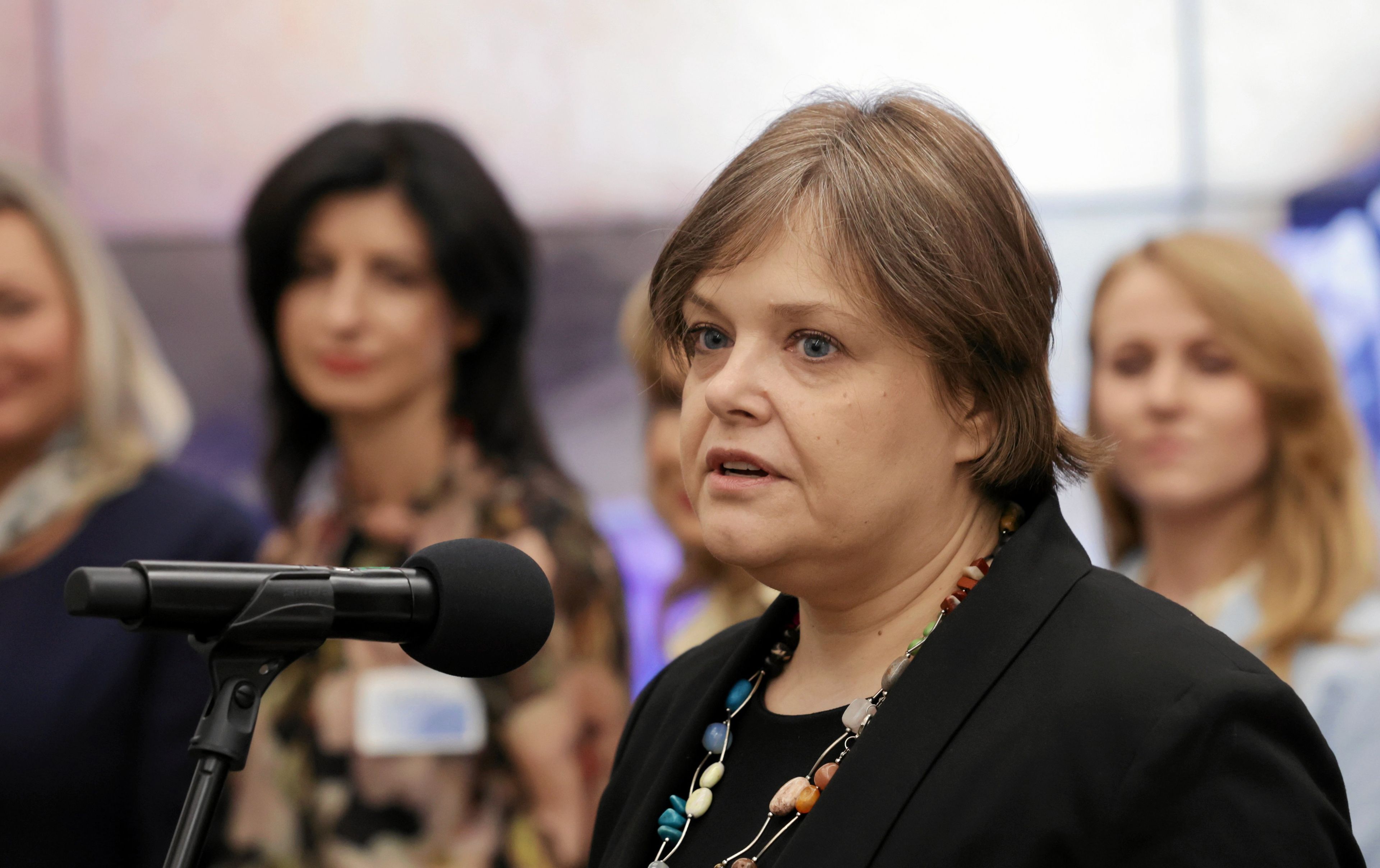 Kobieta w średnim wieku, oficjalnie ubrana, stoi przed mikrofonem. W tle widoczne inne osoby.