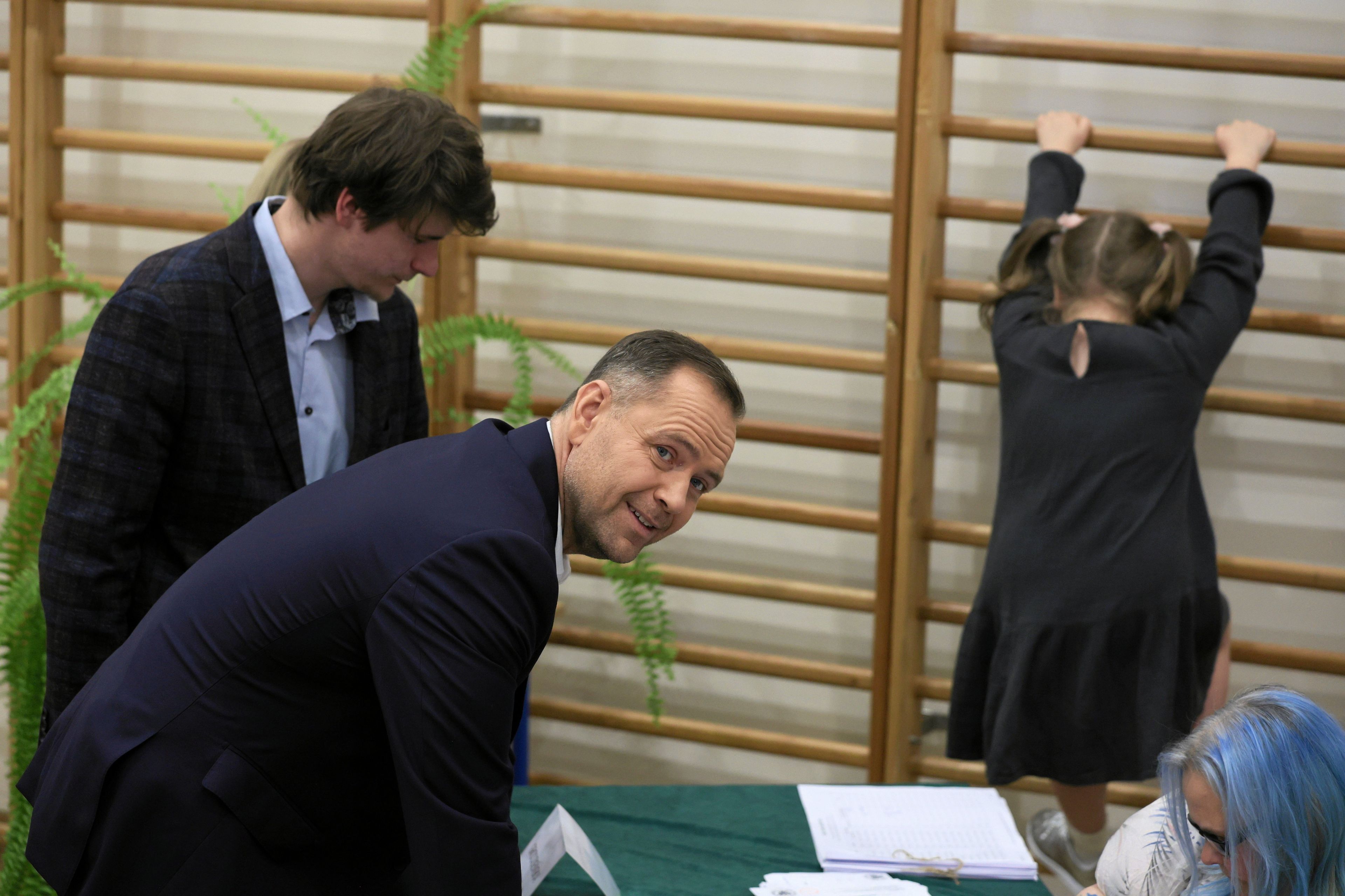 Mężczyzna pochylony nad stołem w lokalu wyborczym, w tle drabinki gimnastyczne, na ktorych bawi się dziewczynka