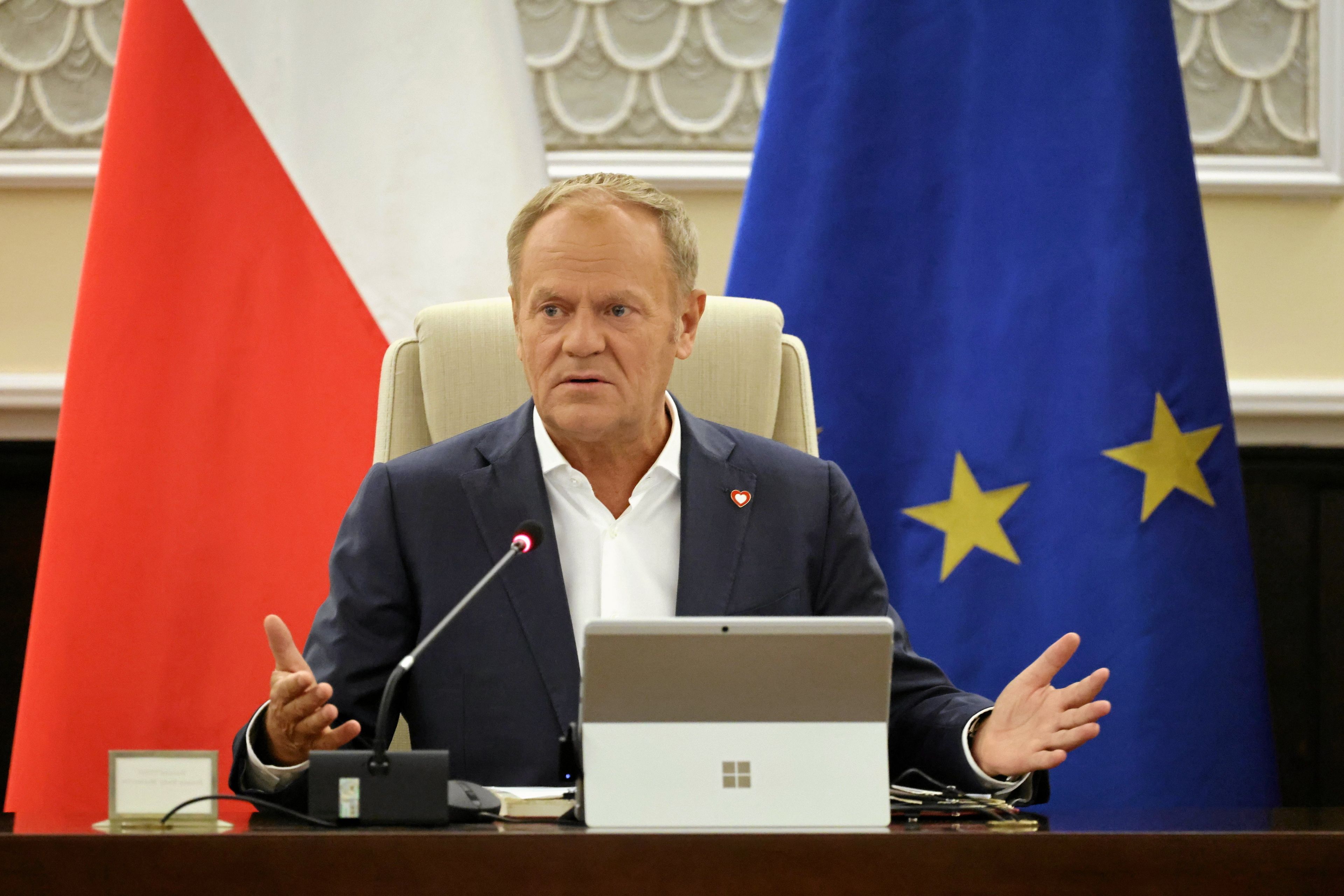 Donald Tusk rozkłada ręce. Przed nim komputer, za nim flagi: biało-czerwona i niebieska z gwiazdkami, europejska