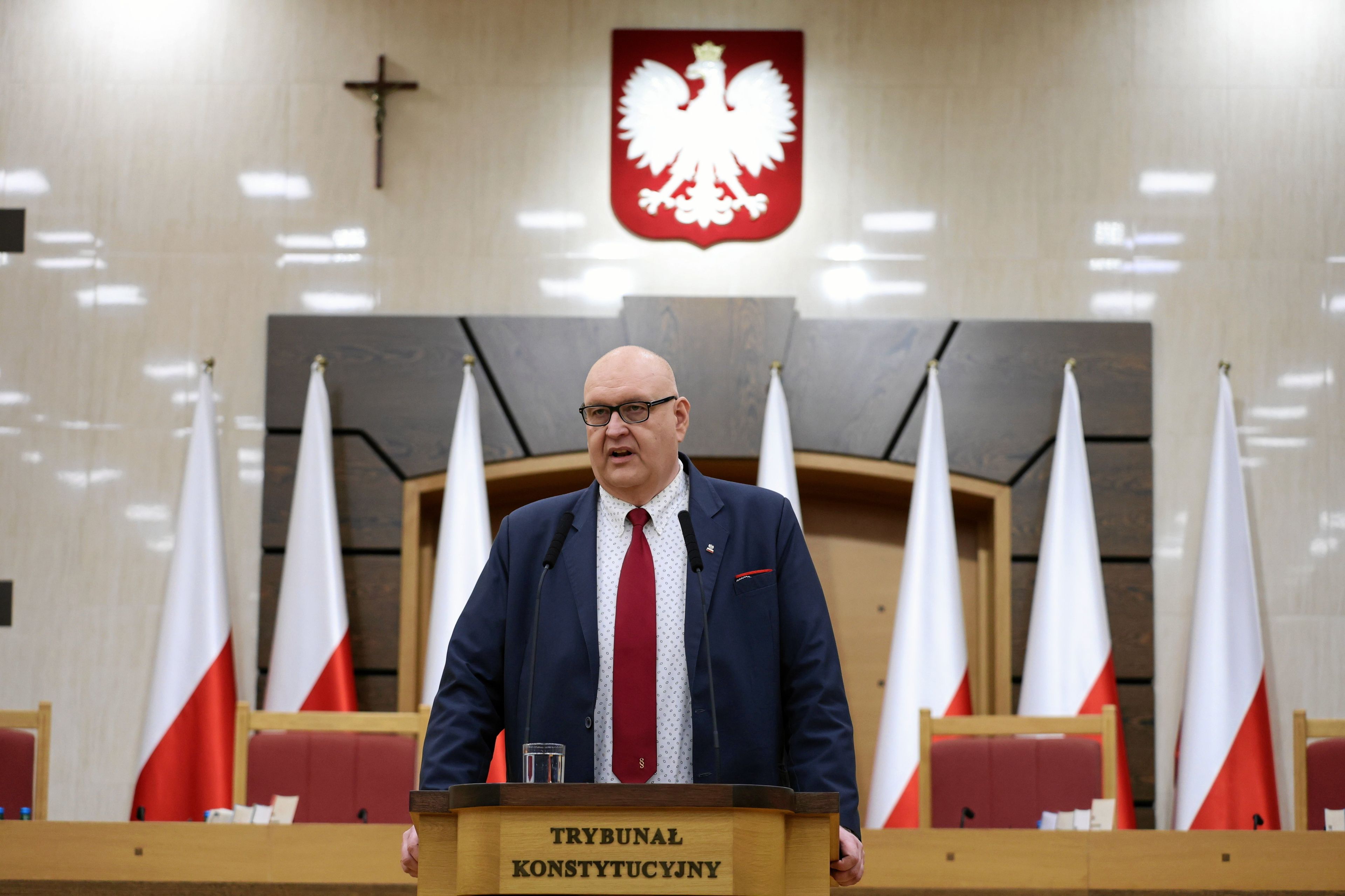 Ogromny męzczyzna w garniturze na mówinicy z napisem Trubunał Konstytucyjny, Z tyłi polskie glagi, godło i kryzż