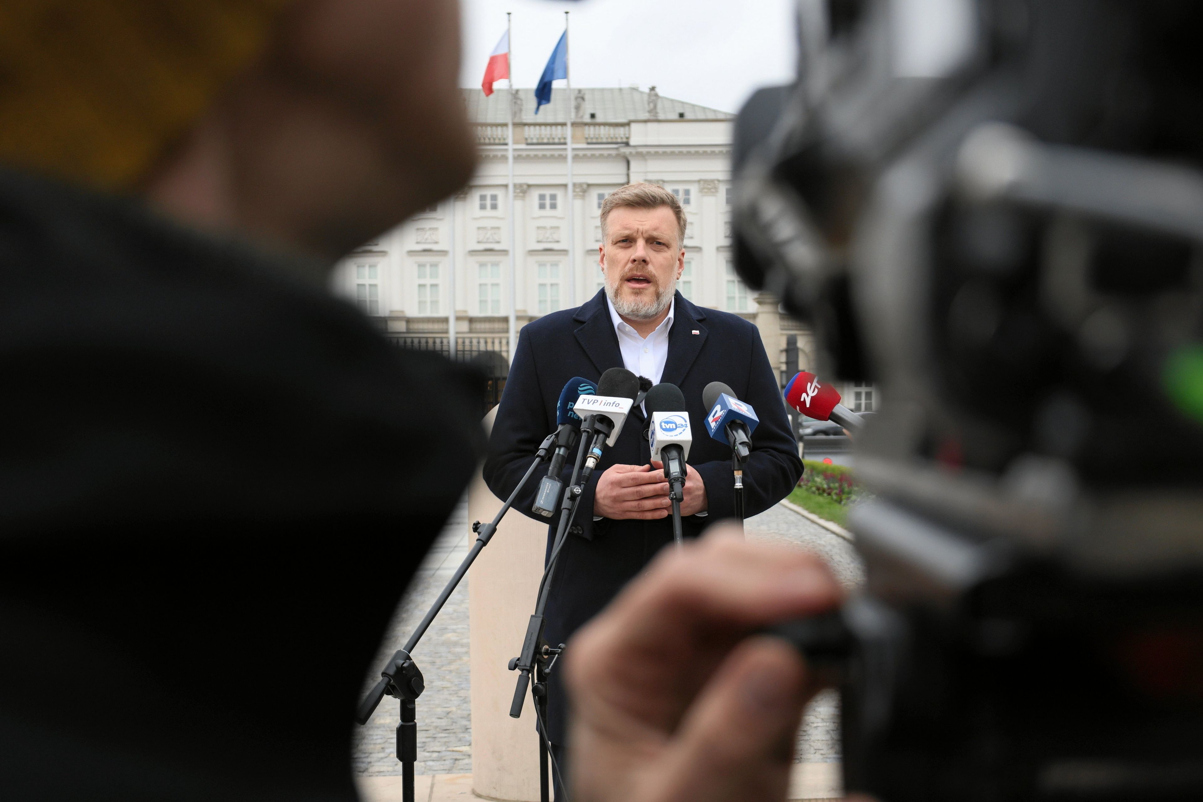 Mężczyzna w płaszczu stoi w tle kadru, zanim Pałac Prezydencki. Na pierwszym planie po obu stronach kamerzyści