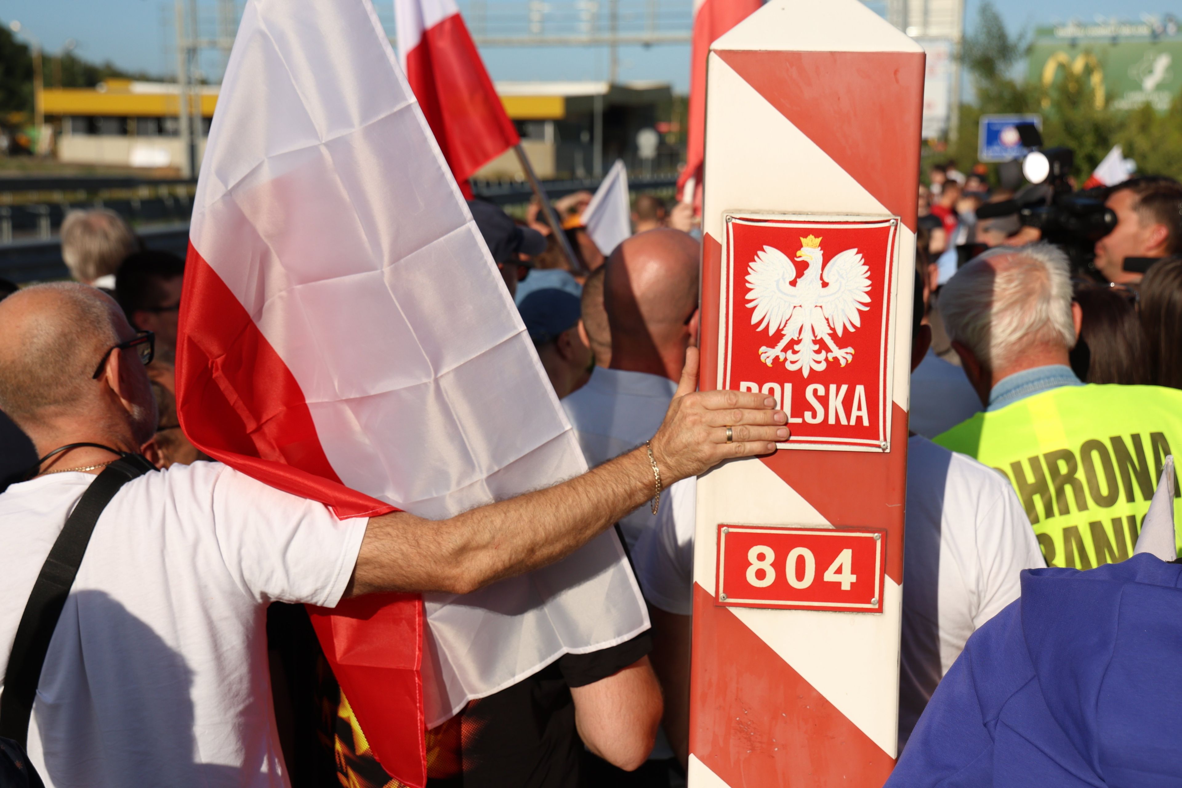Kilka osób z czerwop0-białymi flagami przy słupie granicznym z godłem Polski