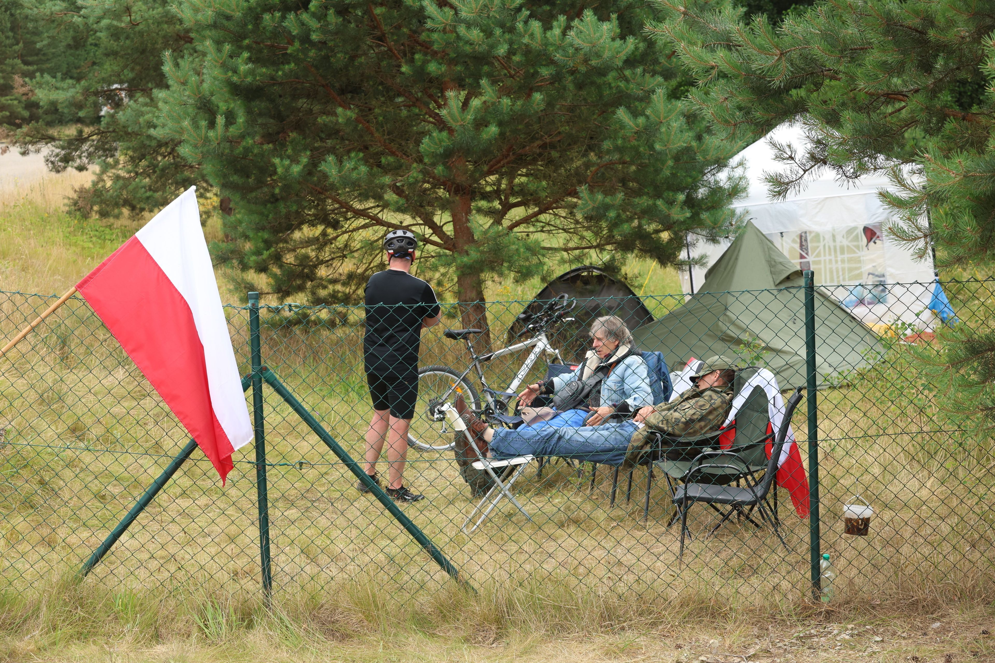 Mężczyźni siedzący na krzesełkach przed namiotami, na pierwszym planie polska flaga.