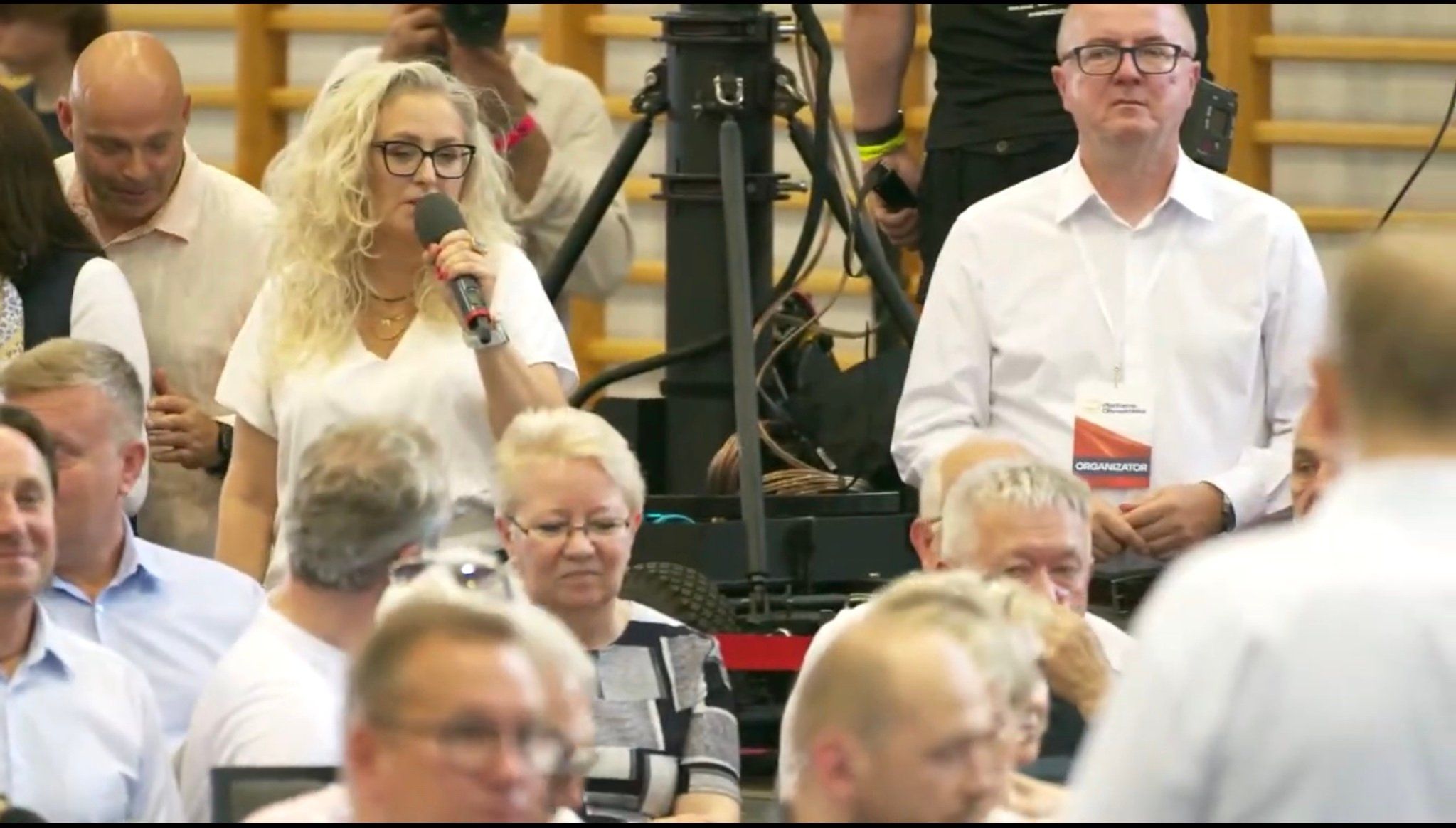Jasnowłosa, otoczona ludxmi kobieta zadaje pytanie