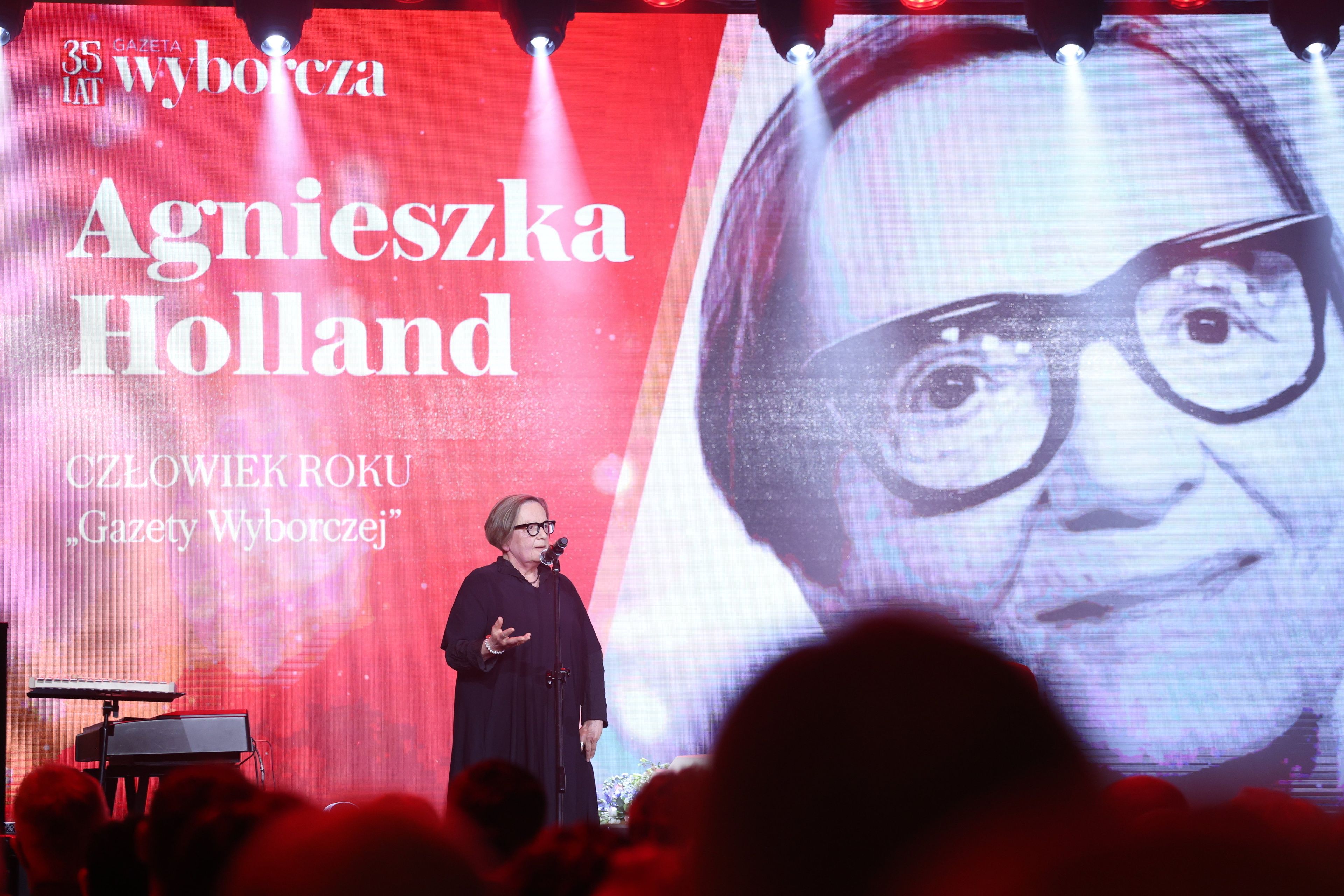 Kobieta w ciemnej sukni z mikrofonem na tle twarzy z napisem Agnieszka Hoollan