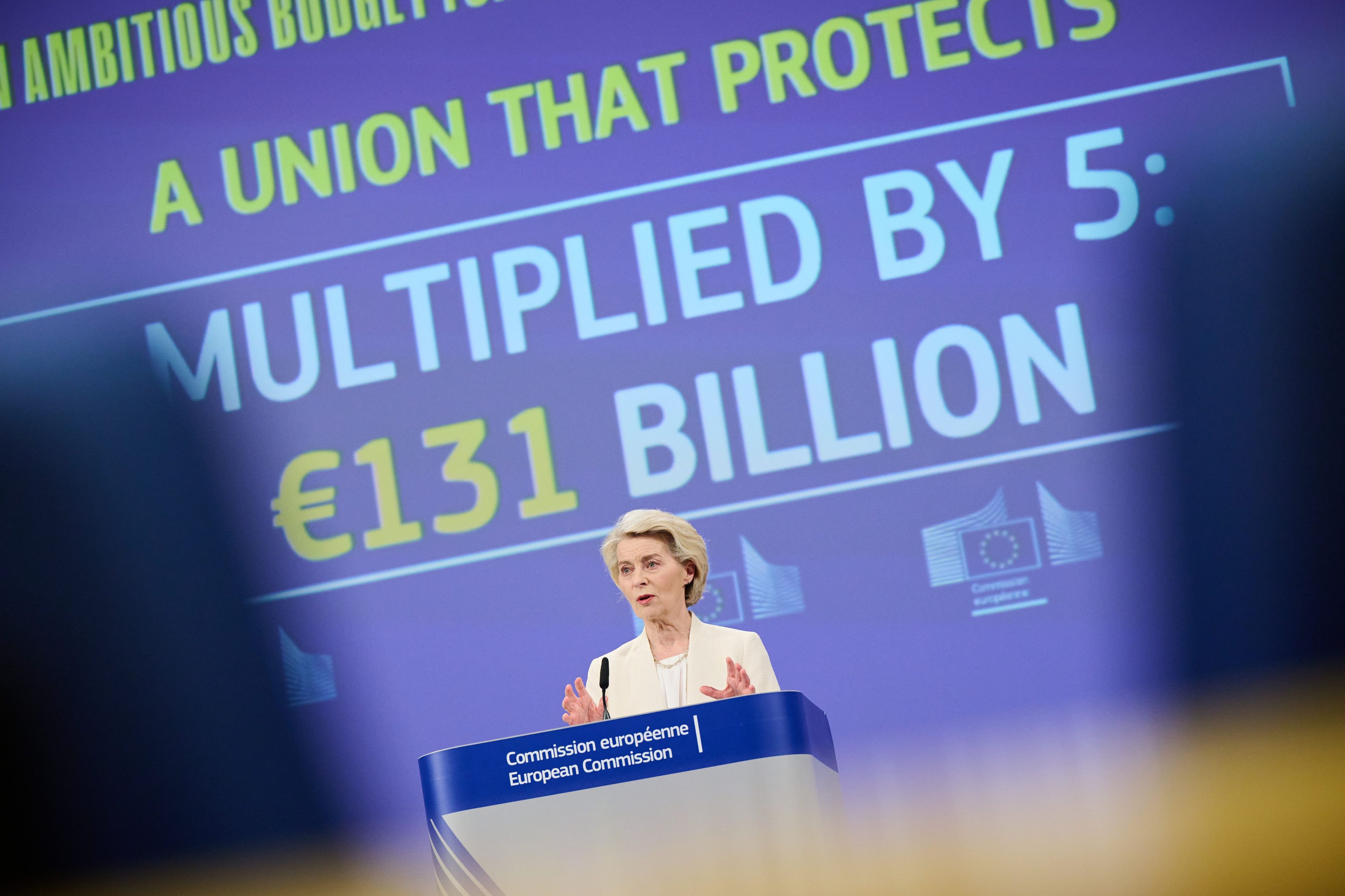 Kobieta o jasnych włosach, ubrana w biały żakiet, przemawia przy mównicy Komisji Europejskiej na tle dużego ekranu z napisem: „Unia, która chroni – zwielokrotnione 5 razy: 131 miliardów euro”.
