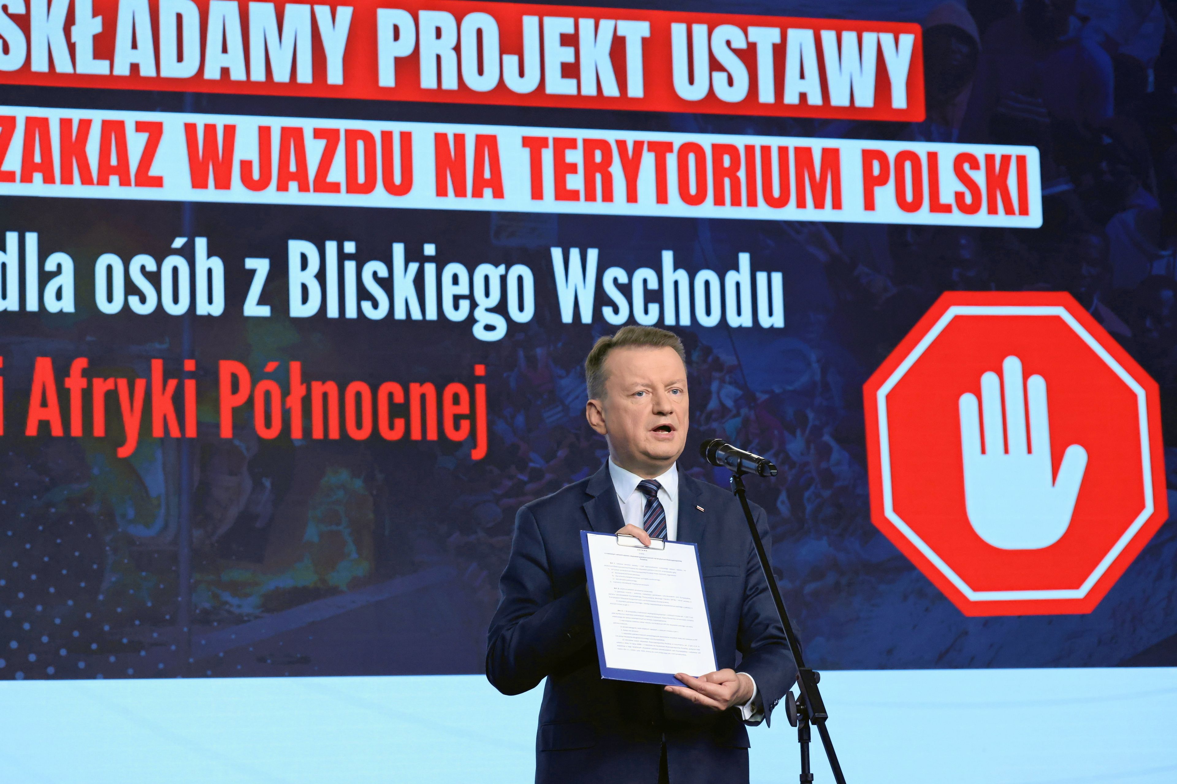 Mariusz Błaszczak podczas konferencji. Pokazuje plik kartek