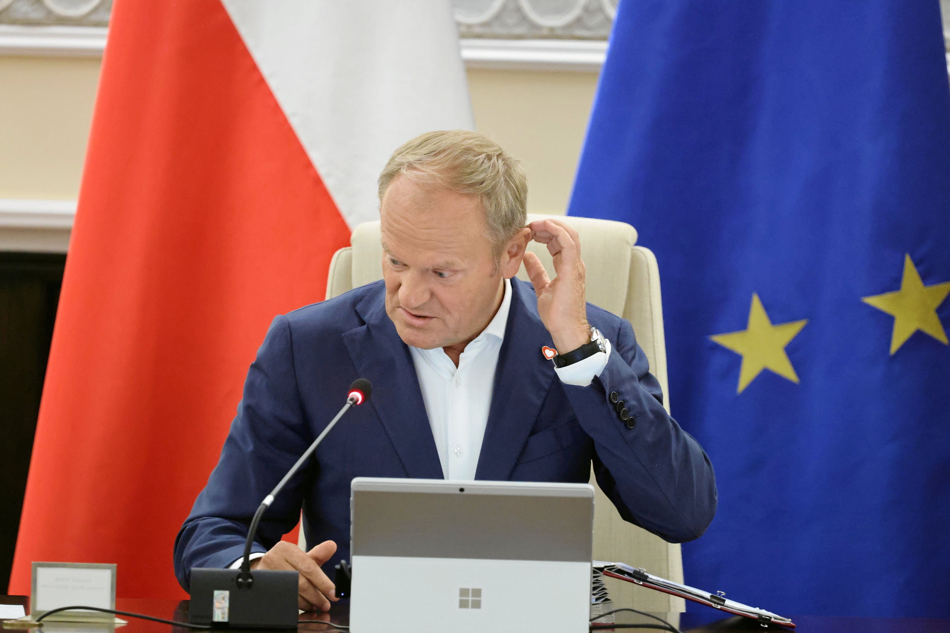 Mężczyzna siedzi, drapie się w głowę, za nim flagi - polska i unijna