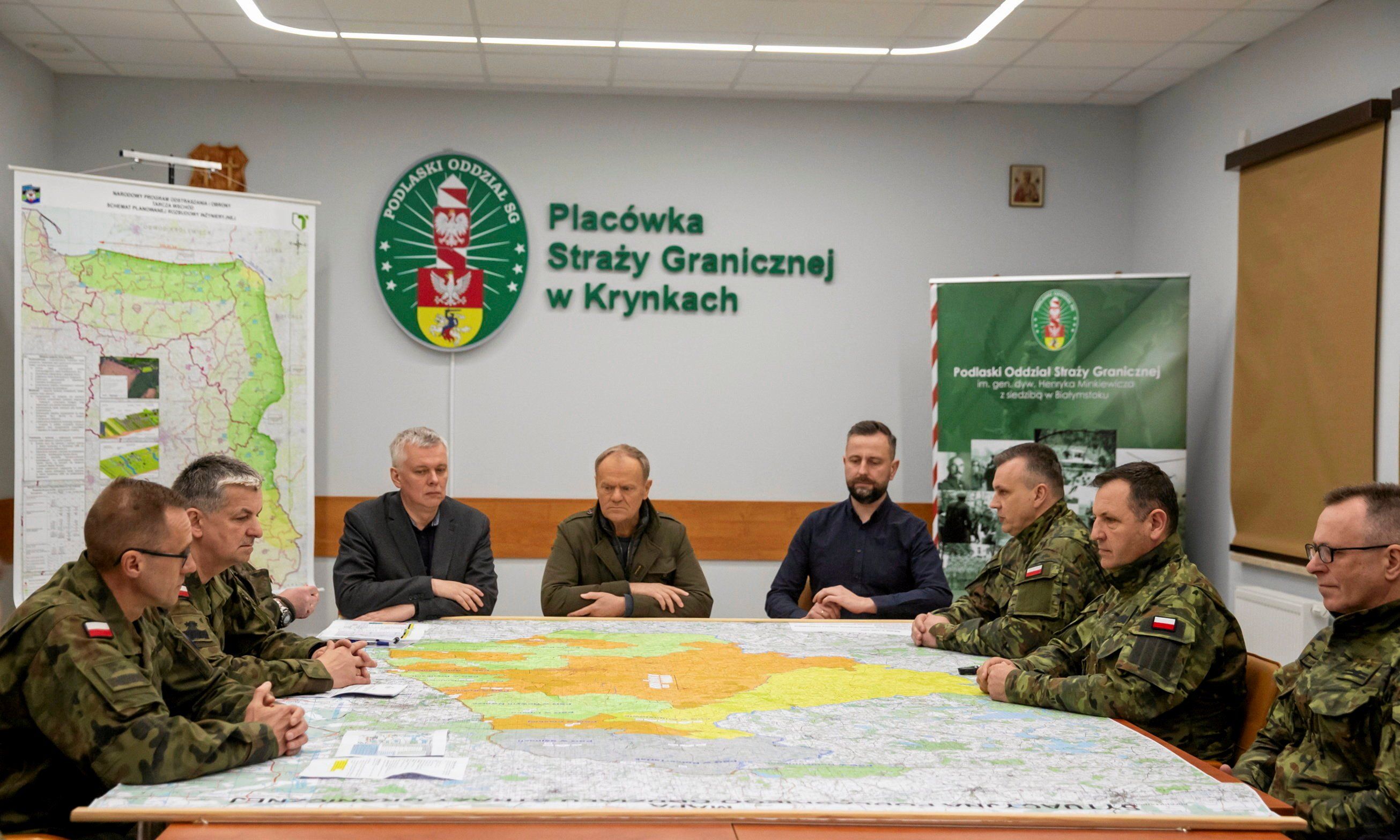 Trzech męzczyzn prezydiuje u stołu, przy którym siedzą ludzie w mundurach. Na ścianie napis "Placówka Straży Granicznej w Krynkavch"