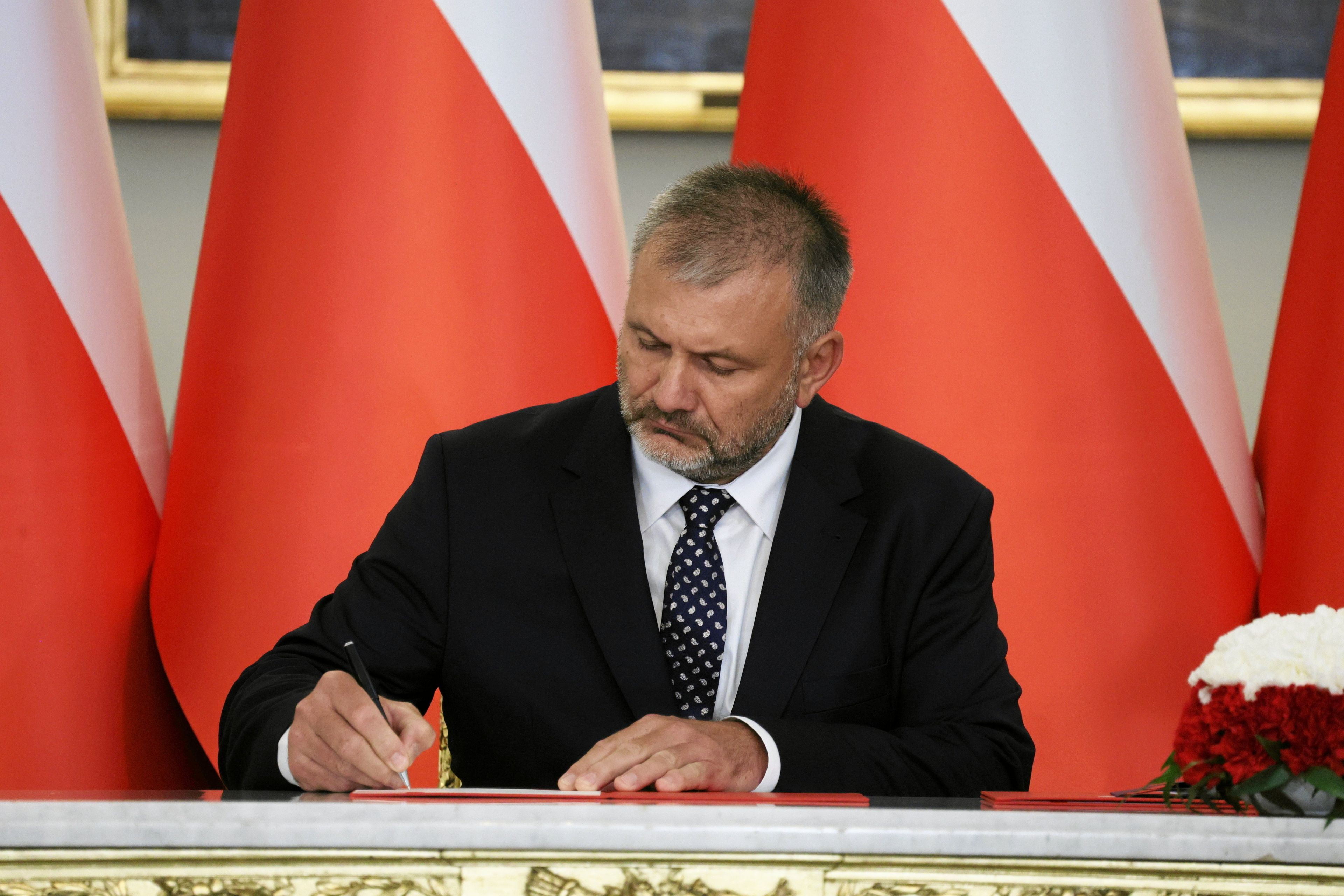 Mężczyzna w garniturze i z zarostem podpisuje dokument. Z tyłu biało-czerwone flagi