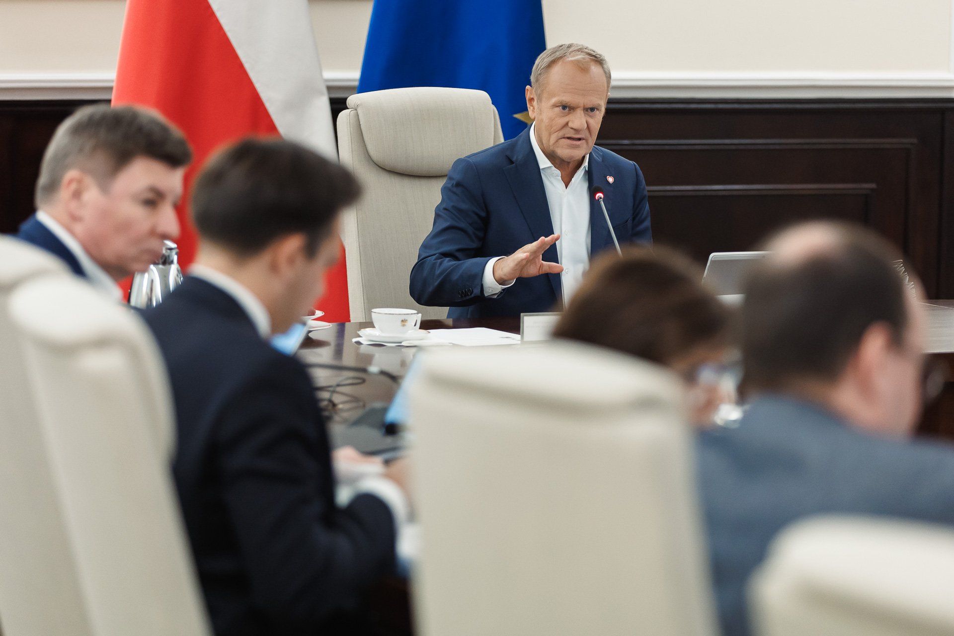Mężczyzna w granatowej marynarce siedzi przy stole konferencyjnym, mówi do mikrofonu i gestykuluje. Wokół niego inni uczestnicy spotkania, filiżanki, dokumenty, flagi Polski i UE w tle.