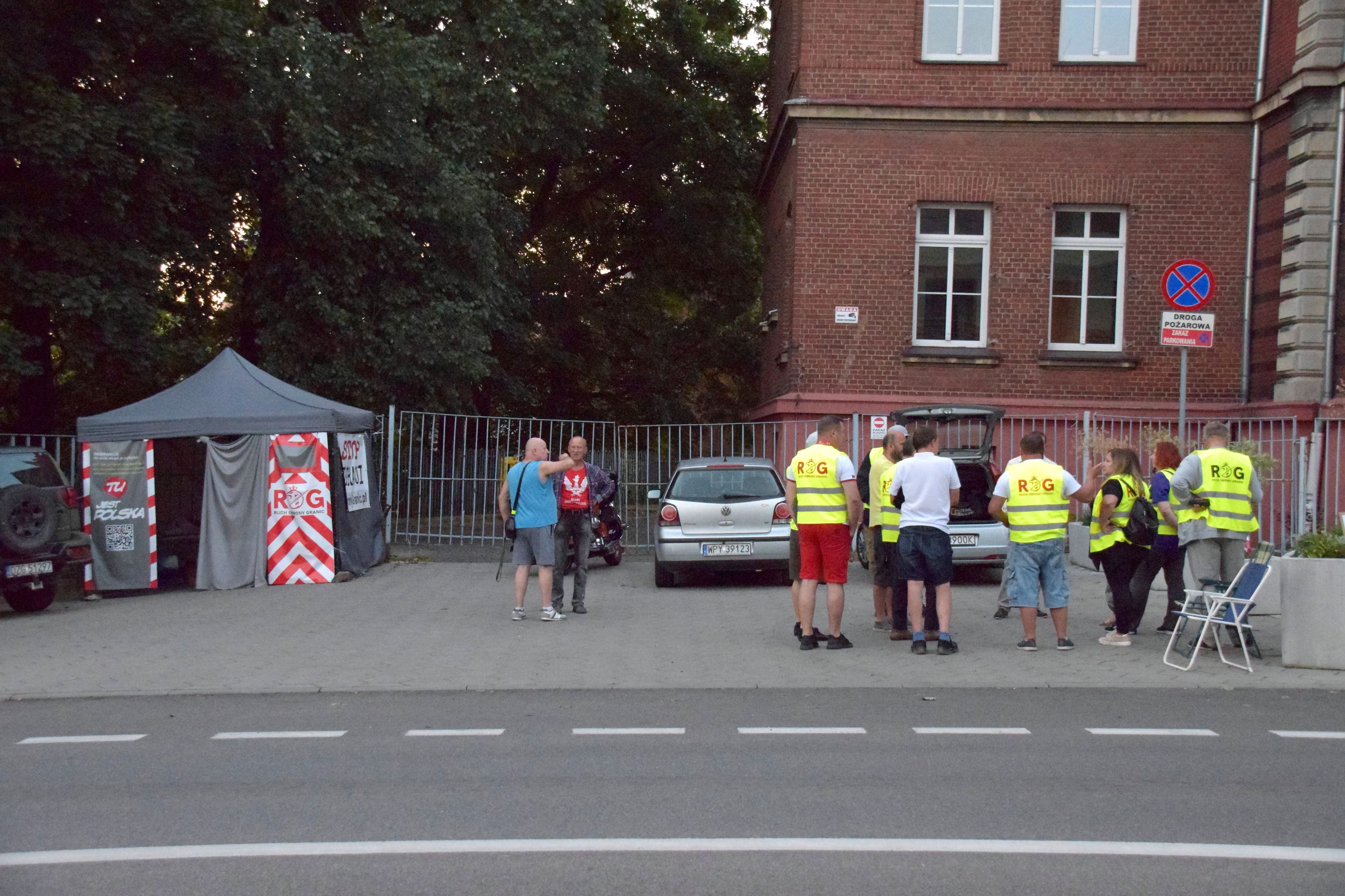 Grupa osób w kamizelkach odblaskowych z napisem „R.O.G.” stoi przed budynkiem z czerwonej cegły; obok namiot z banerami i napisem „STOP IMIGRACJI”, w tle ogrodzenie i drzewa.