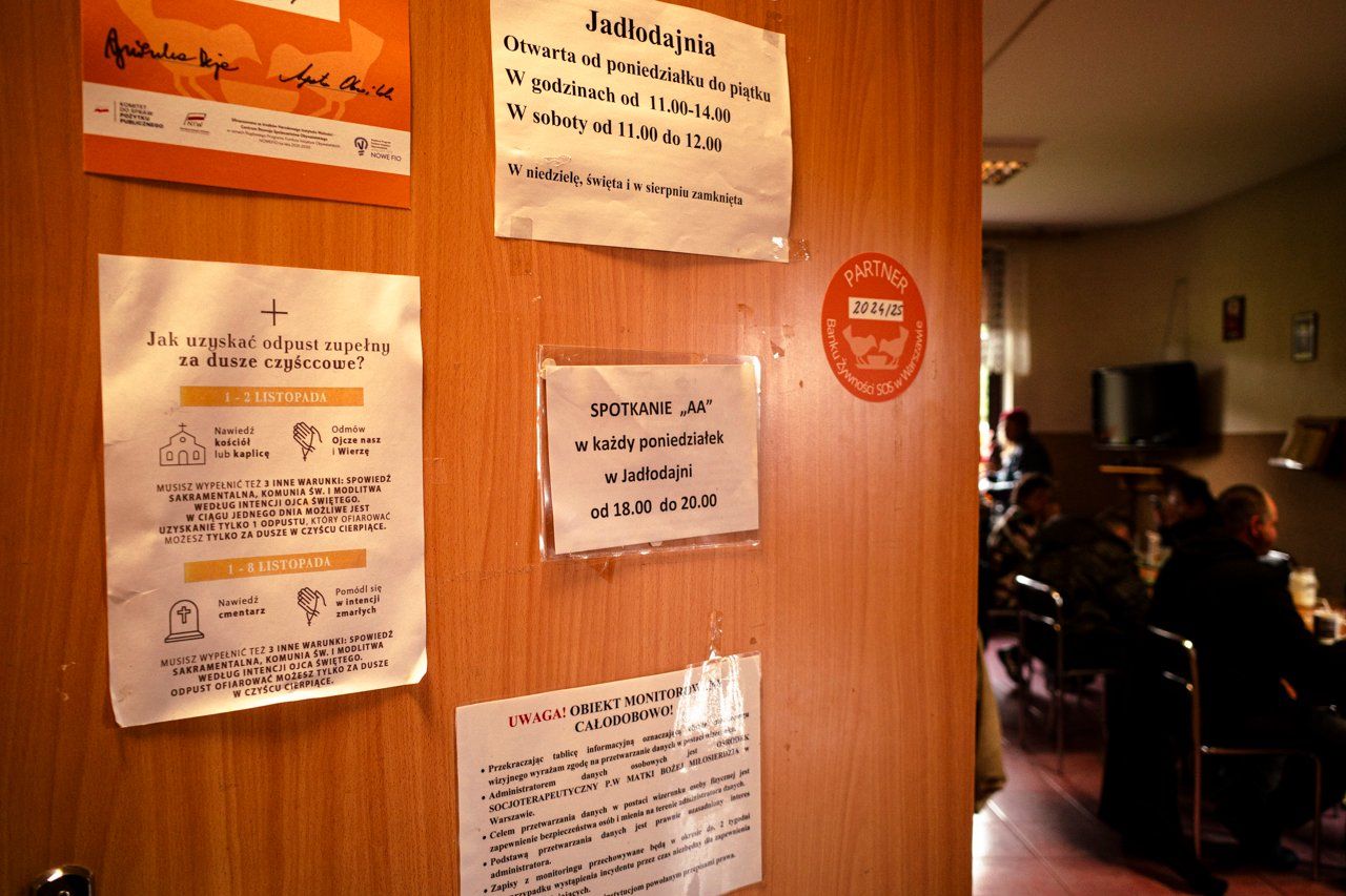 Fotografia. Uchylone drzwi z różnymi informacjami, w głębi widać siedzące osoby.