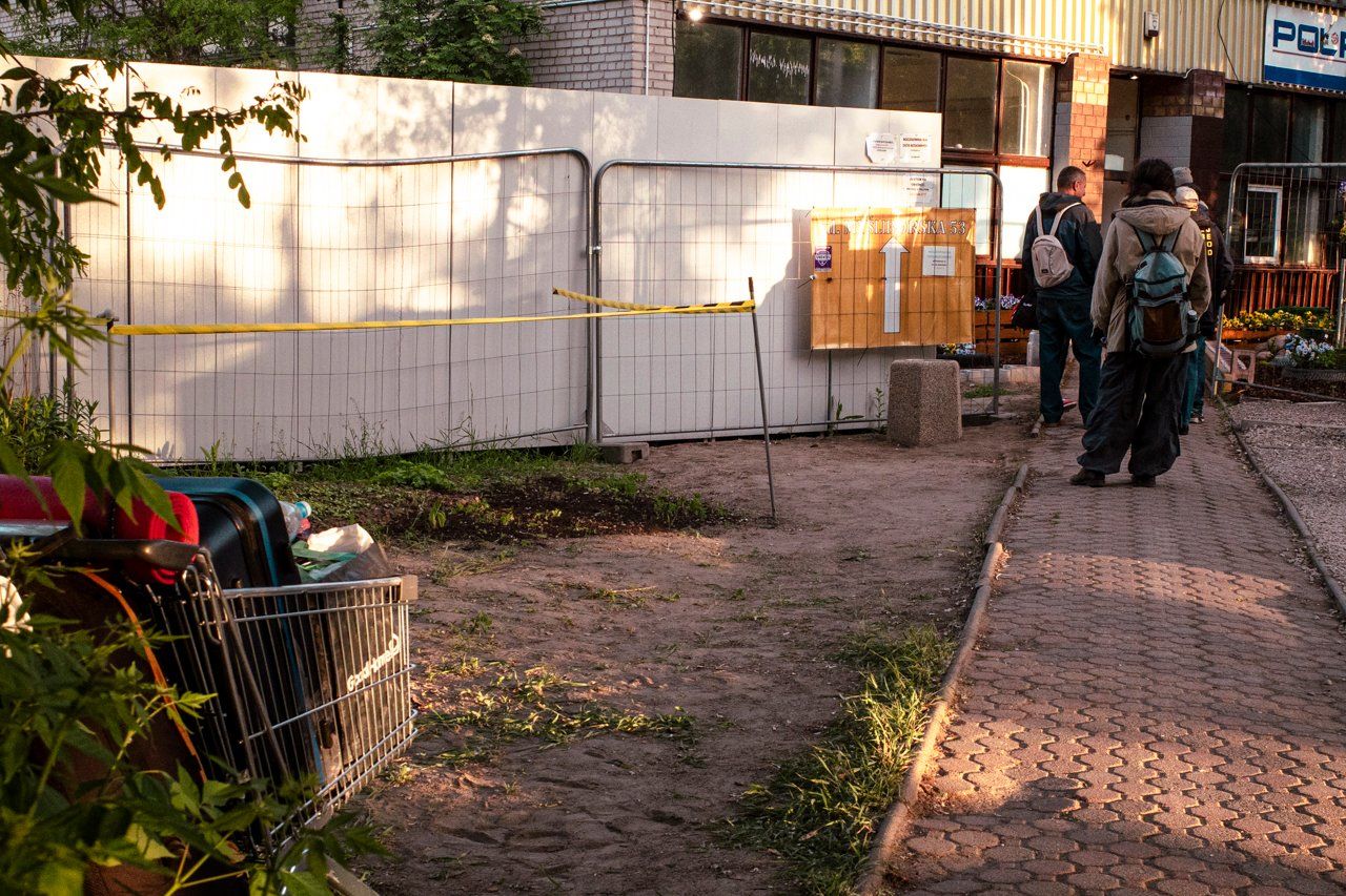 Wejście do parterowego, prowizoryczne ogrodzenie, przed drzwiami stoją osoby z plecakami
