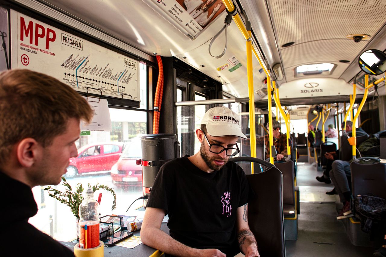 Wnętrze miejskiego autobusu, przy oknie, przy stoliku siedzą dwaj młodzi ludzie. Nad nimi biała tablica z napisem MPP.dzą