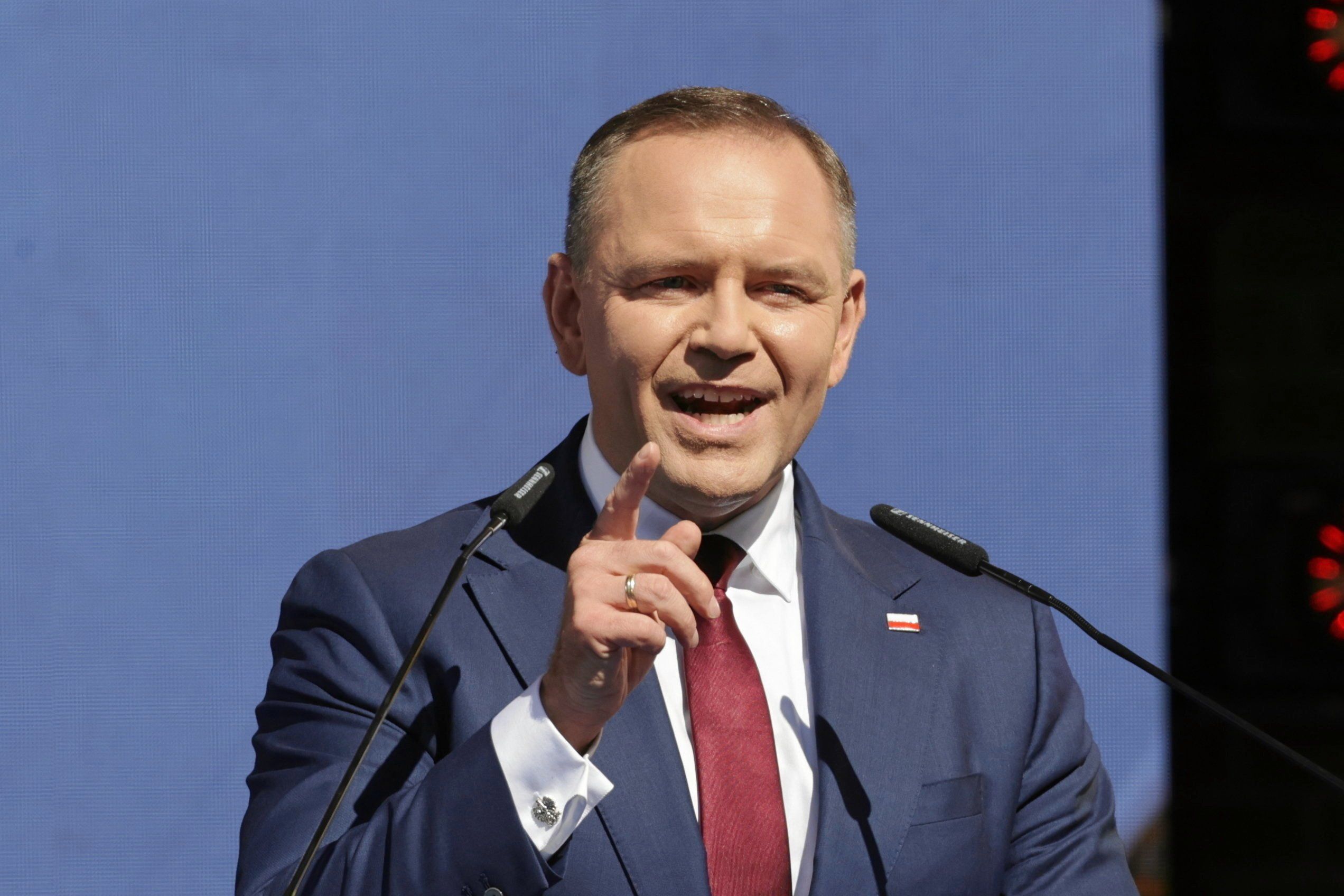 Mężczyzna w niebieskawym garniturze i czerwonym krawacie przemawia machając ręką