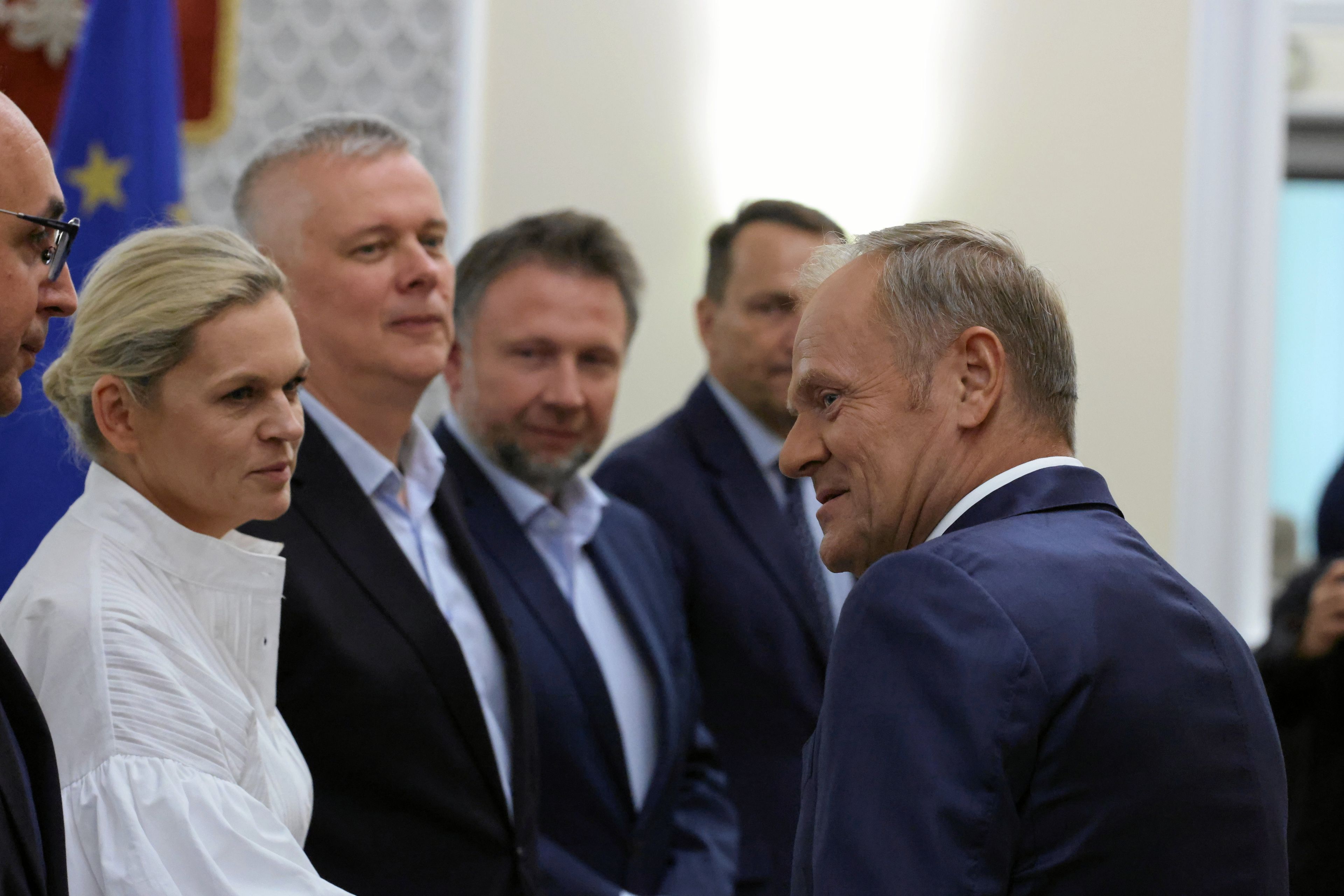 Kobieta w białym kostiumie wita się z mężczyzną w ciemnym garniturze, wokół mężczyźni w garniturach