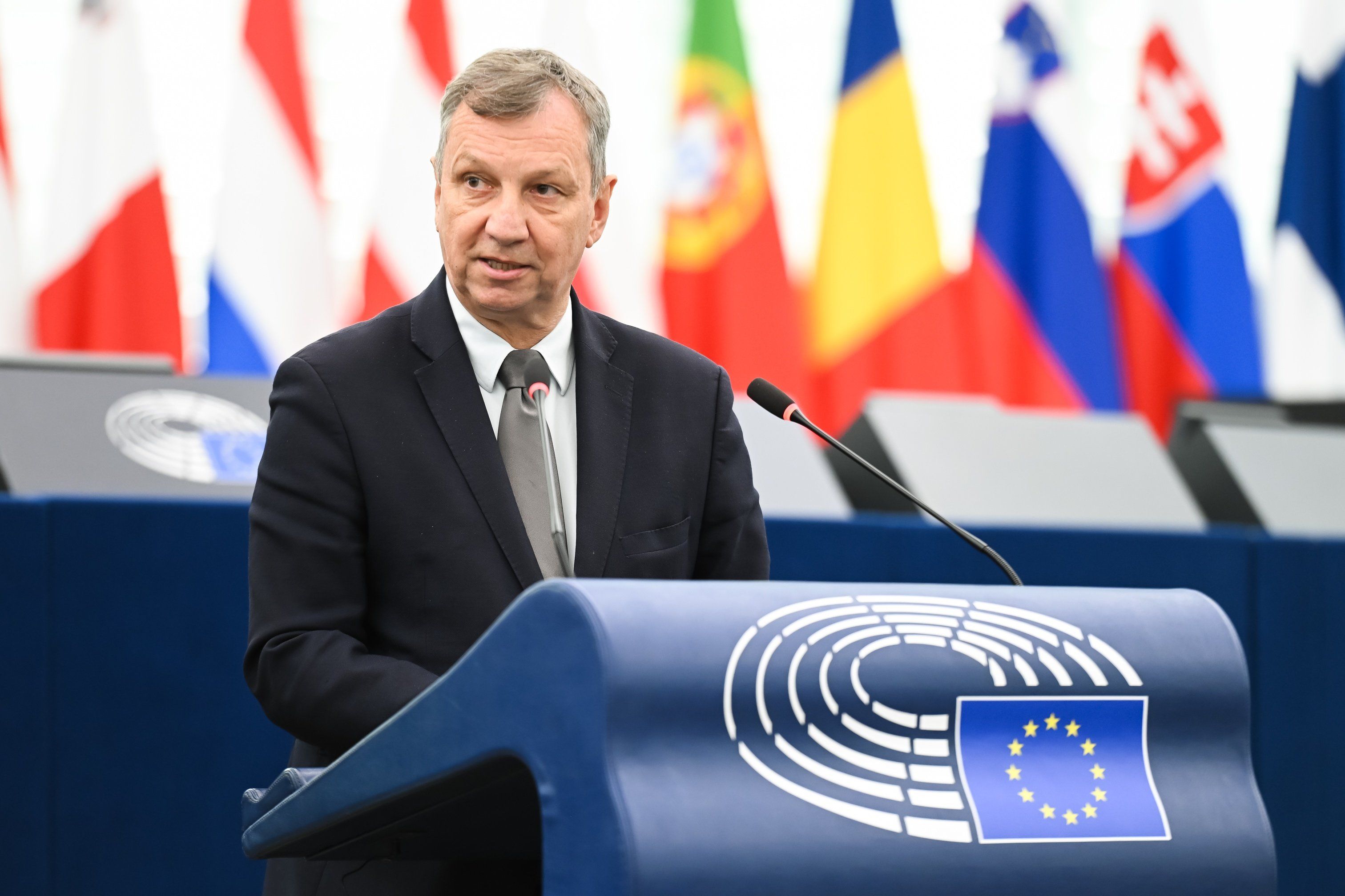 Mężczyzna w garniturze przemawia z mównicy Parlamentu Europejskiego. Za nim widać flagi państw UE i logo Unii Europejskiej. Na sali obecni są inni uczestnicy obrad.