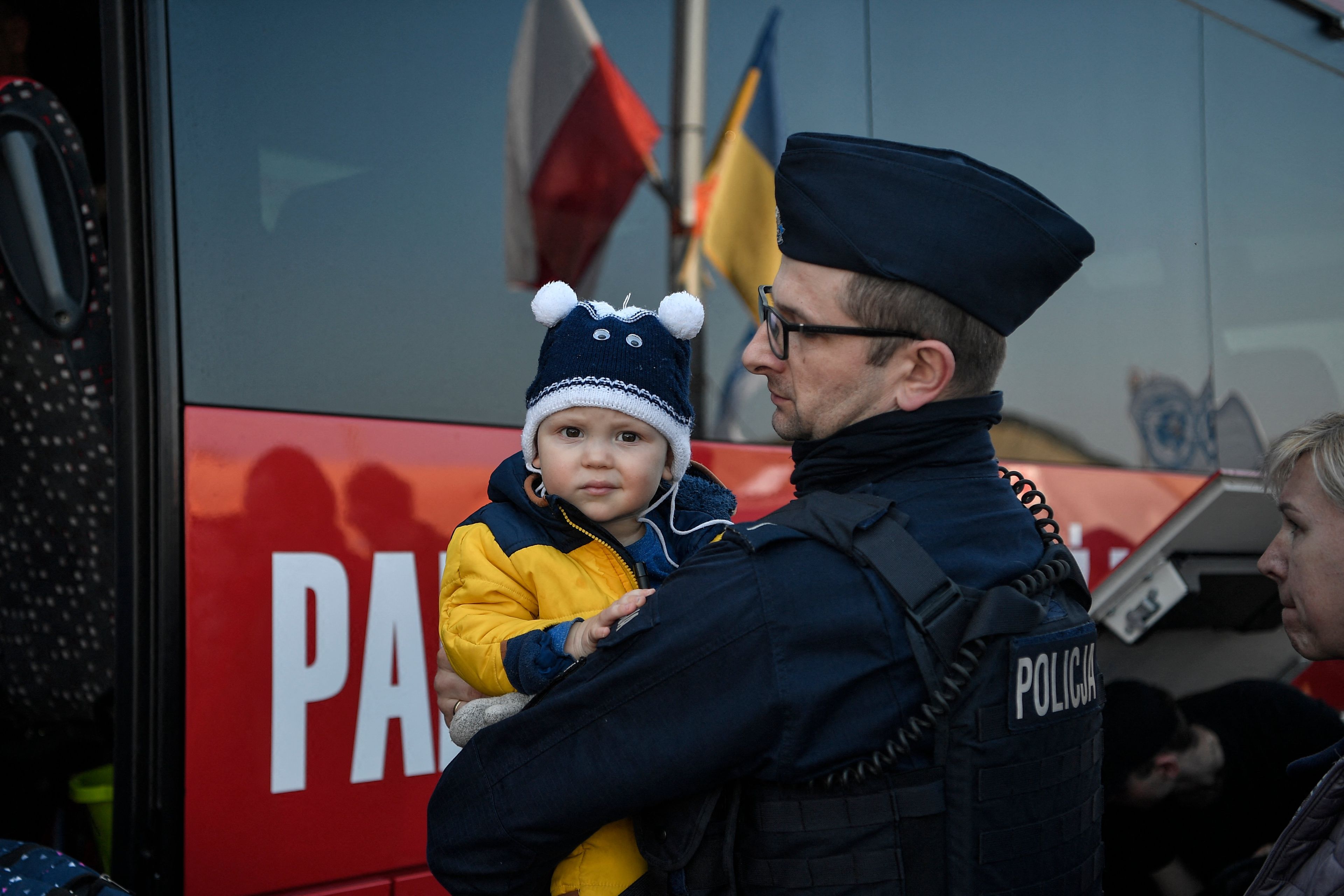 Na zdjęciu funkcjonariusz policji trzyma małe dziecko. W tle widać flagi biało-czerwoną i niebiesko-żółtą