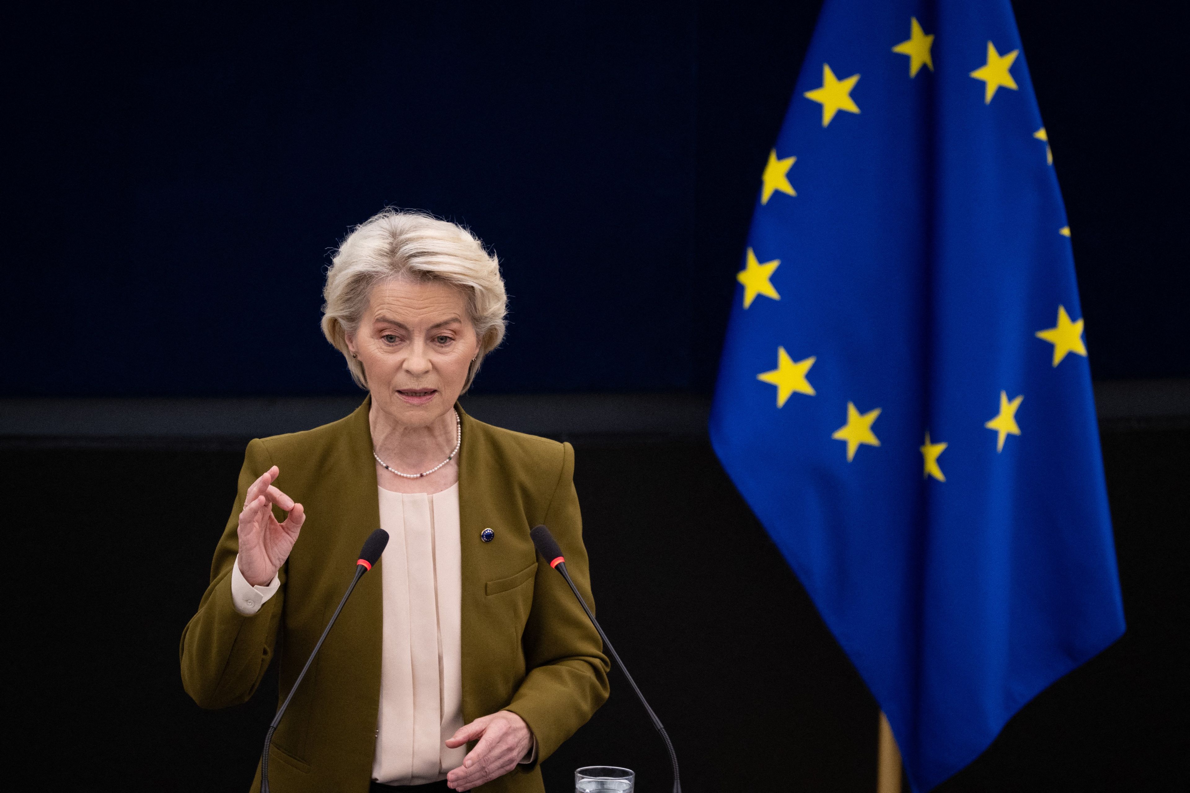 Elegancka kobieta w beżowej marynarce przemawia na tle 12 gwiazdek Unii Europejskiej