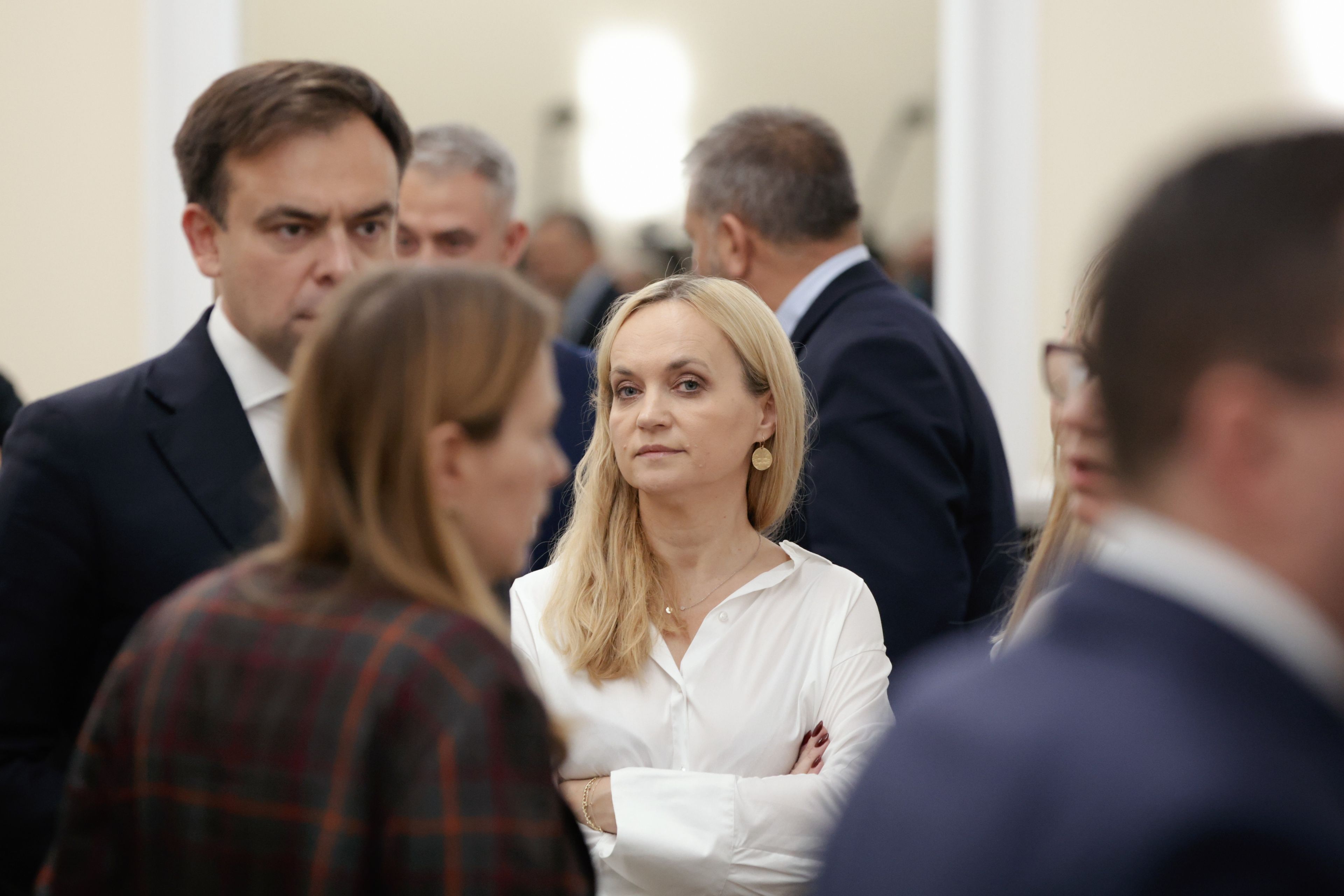 Jasnowłosa kobieta w białej bluzce w otoczeniu ludzi w ciemnych marynarkach