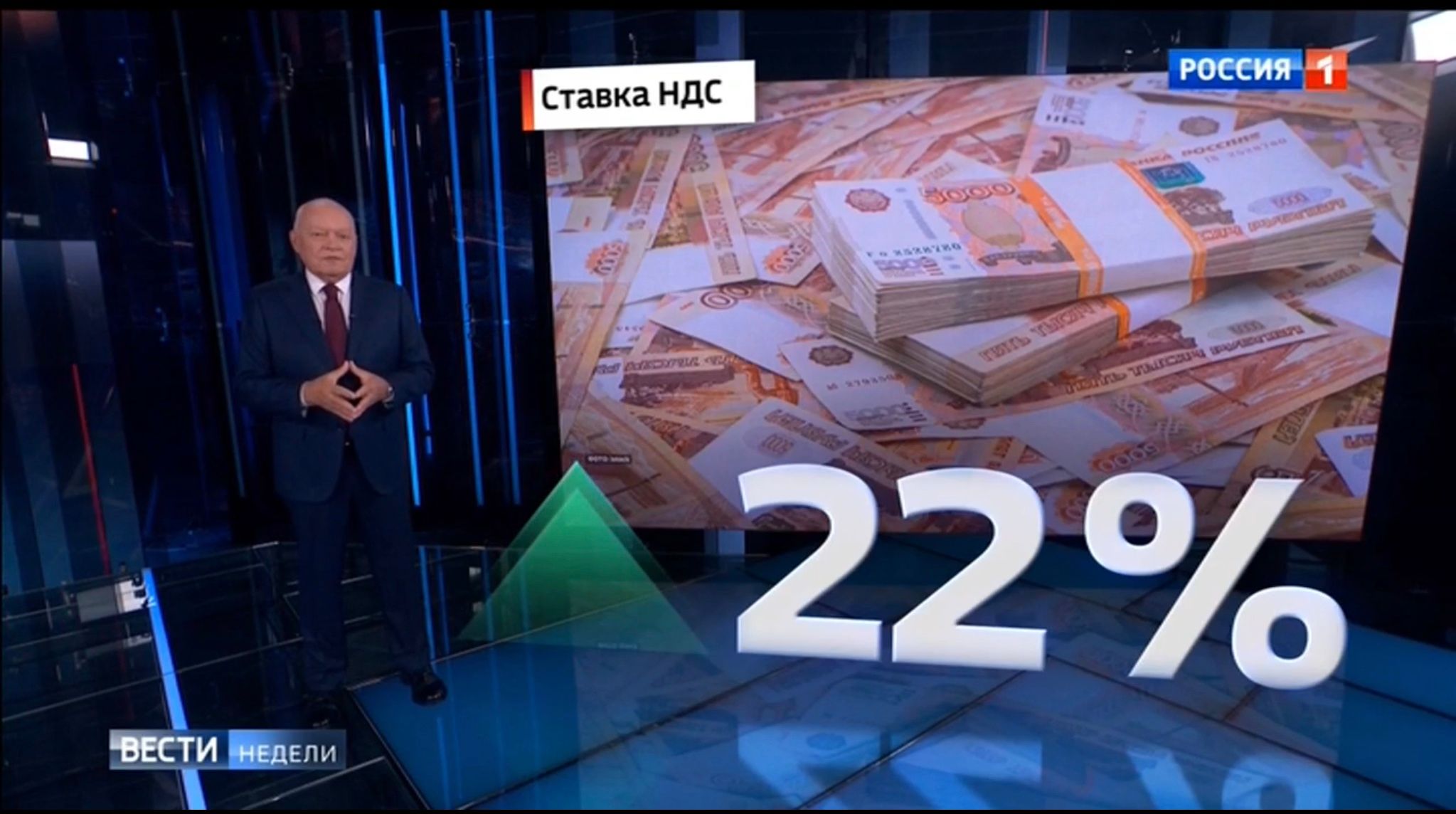 Starszy męzczyzna ze złozonymi w kozyczek dlońmi stoi na tle ekranu z pieniędzmi i wielkim napisem "22%"
