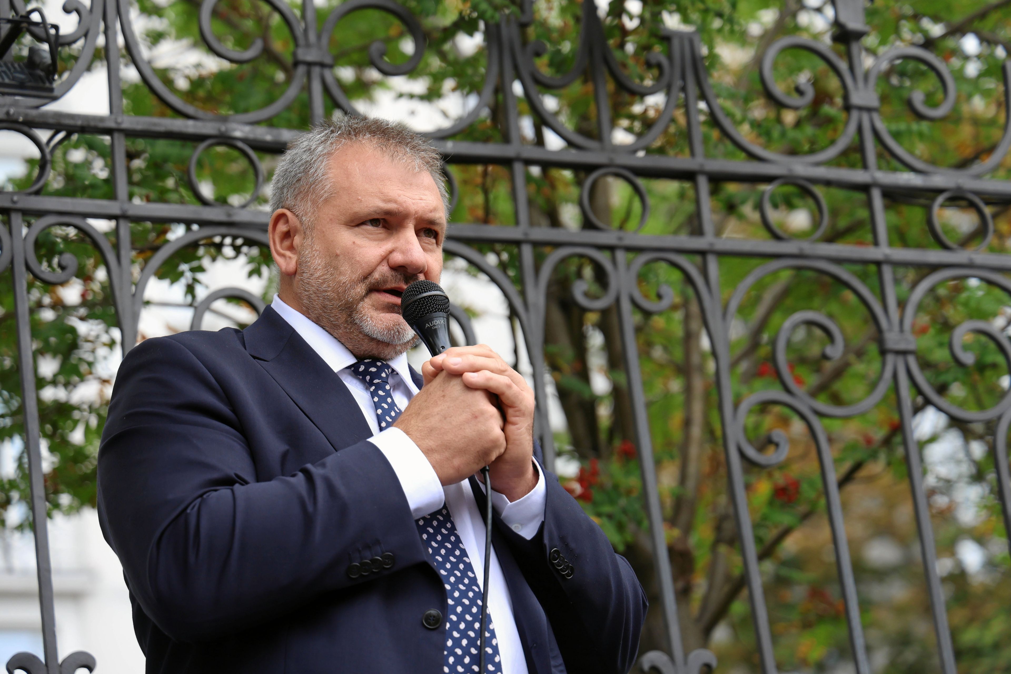 Krótko ostrzyżony mężczyzna w średnim wieku z mikrofonem w ręku stoi na tle metalowego ogrodzenia