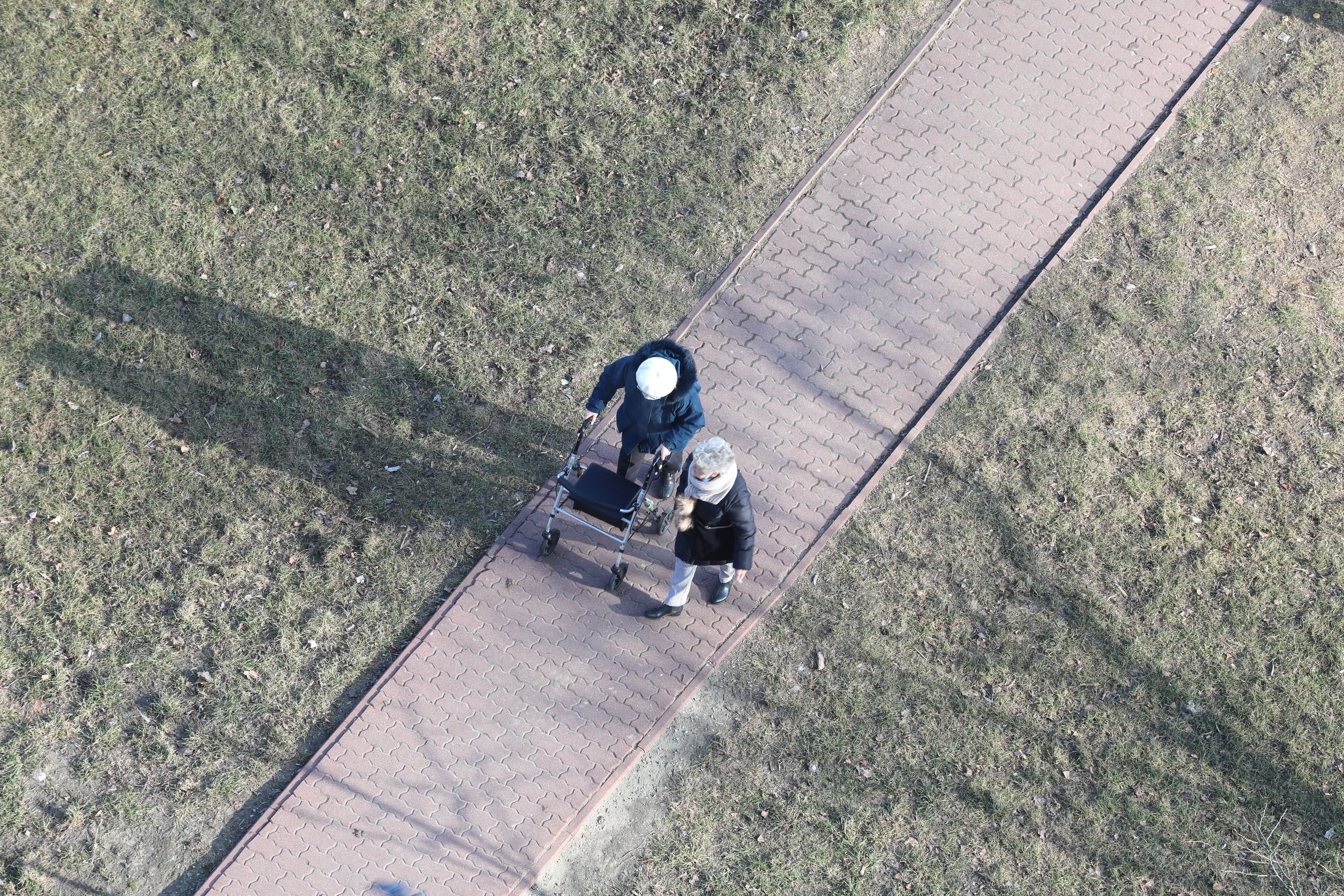 Starsza kobieta idzie z pomocą chodzika. Obok młodsza osoba. WIdok z góry