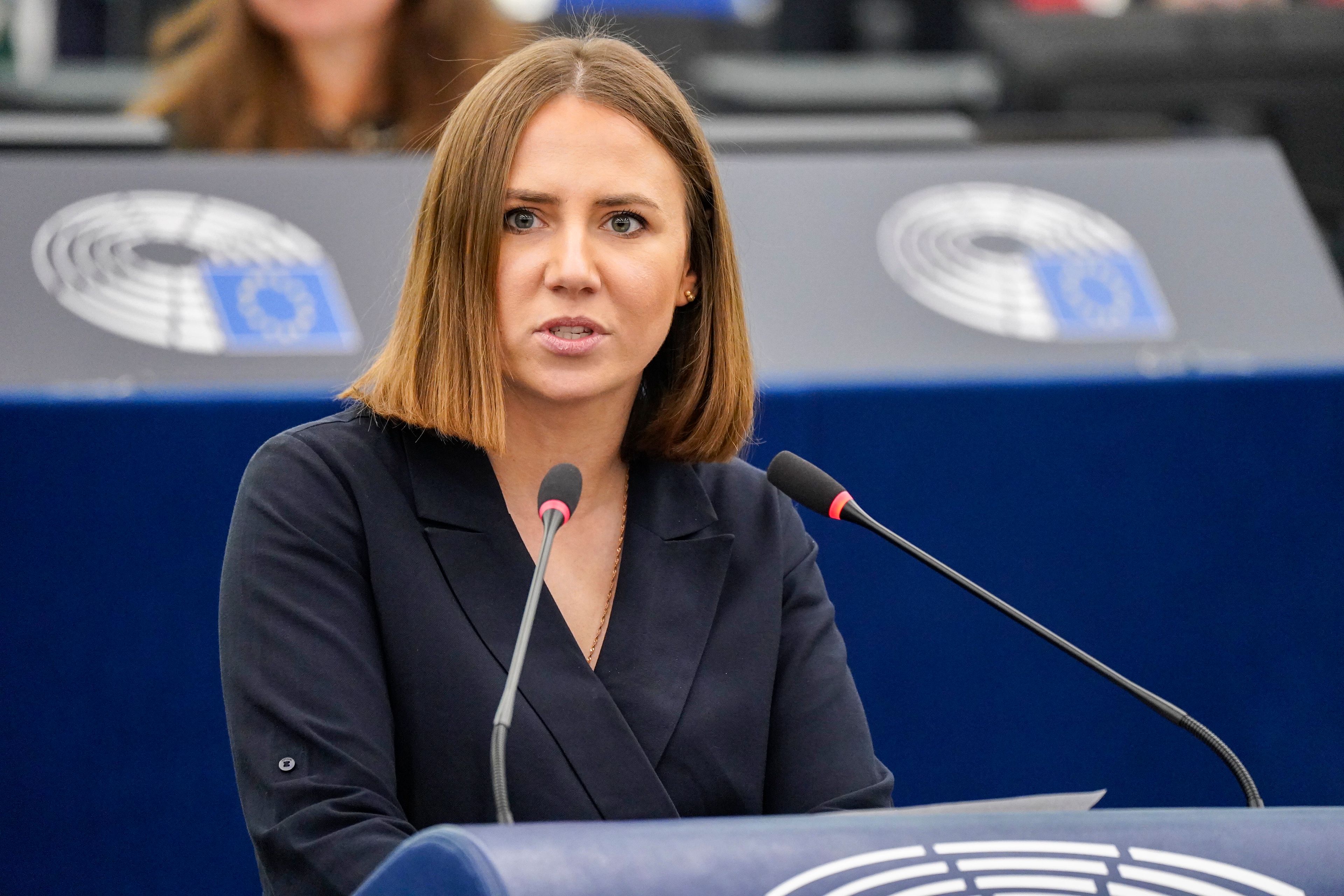 Młoda kobieta z krótkimi brązowymi włosami w ciemnej, kopertowej sukience przemawia z mównicy Parlamentu Europejskiego, w tle widać nieostre logo PE