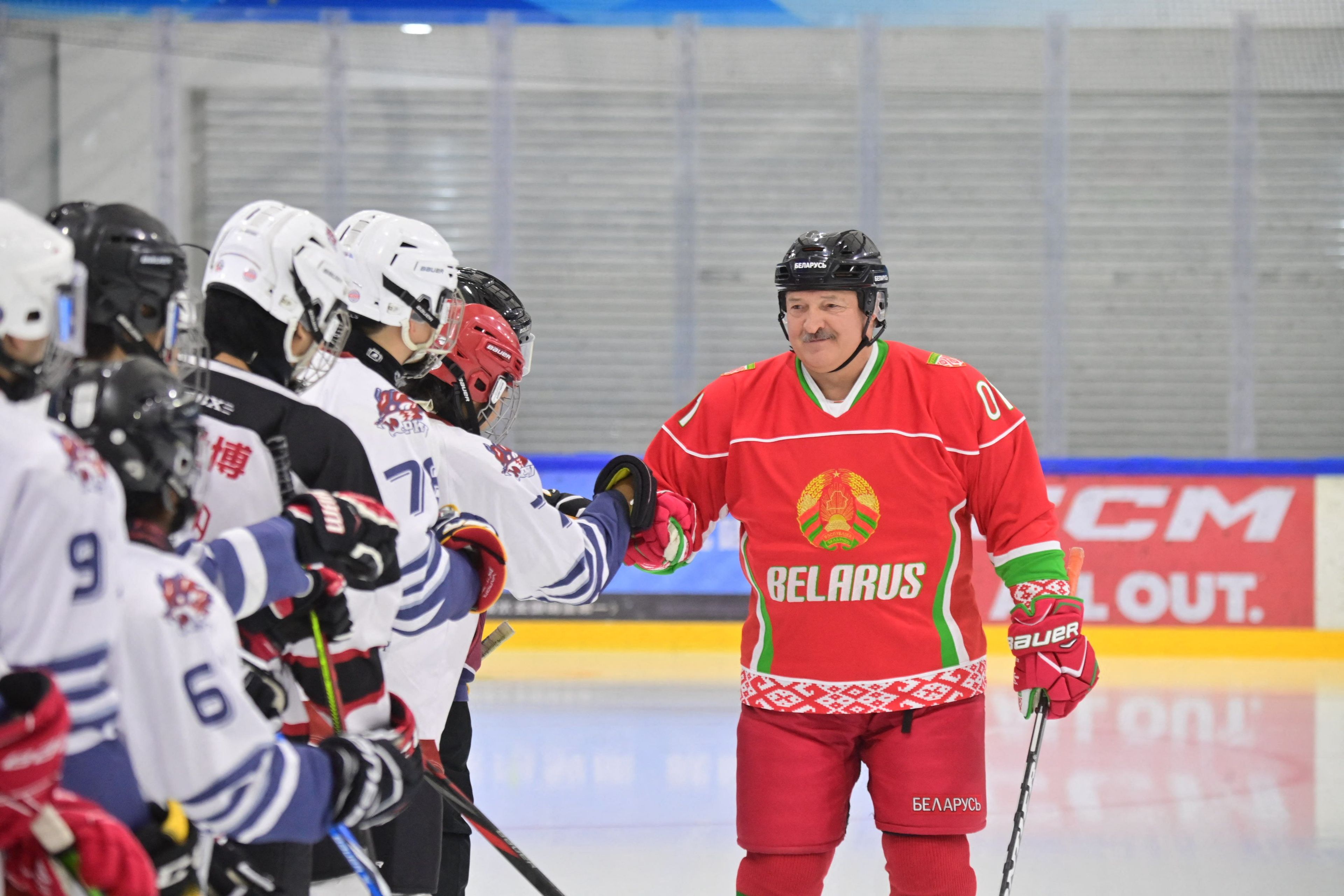 Masywny mężczyzna z wąsem w stroju hokejowym z napisem Belarus na czerwonej bluzie wita się z hokeistami w białych strojach.
