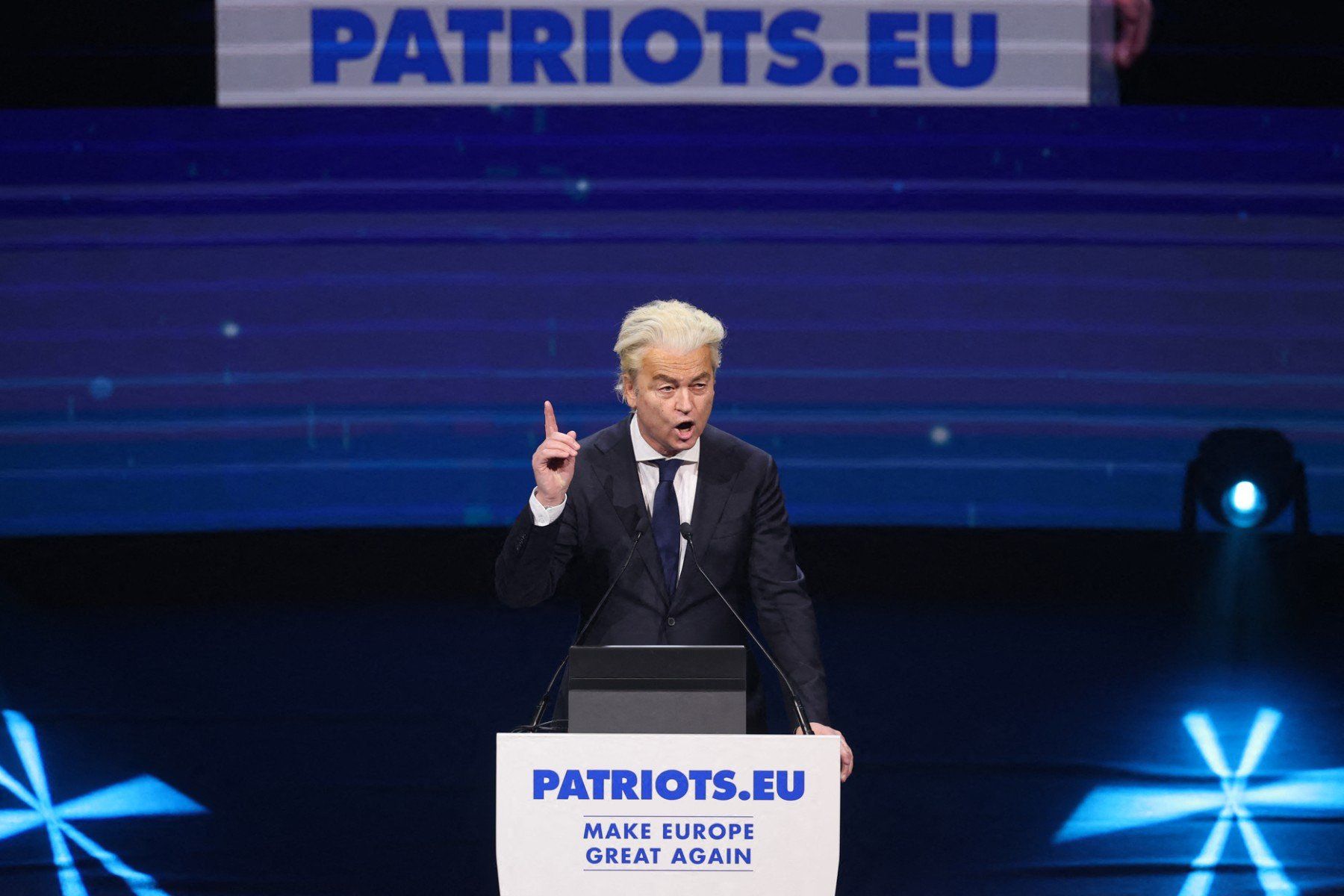 Mężczyzna z tlenionymi blond włosami w czarnym garniturze stoi przy mównicy na scenie, przemawia energicznie, ma uniesiony do góry palec wskazujący prawej ręki, jakby coś z zaangażowaniem wyjaśniał. Tło jest ciemnogranatowe, w tle widać kawałek baneru z napisem "Patrioci dla Europy"