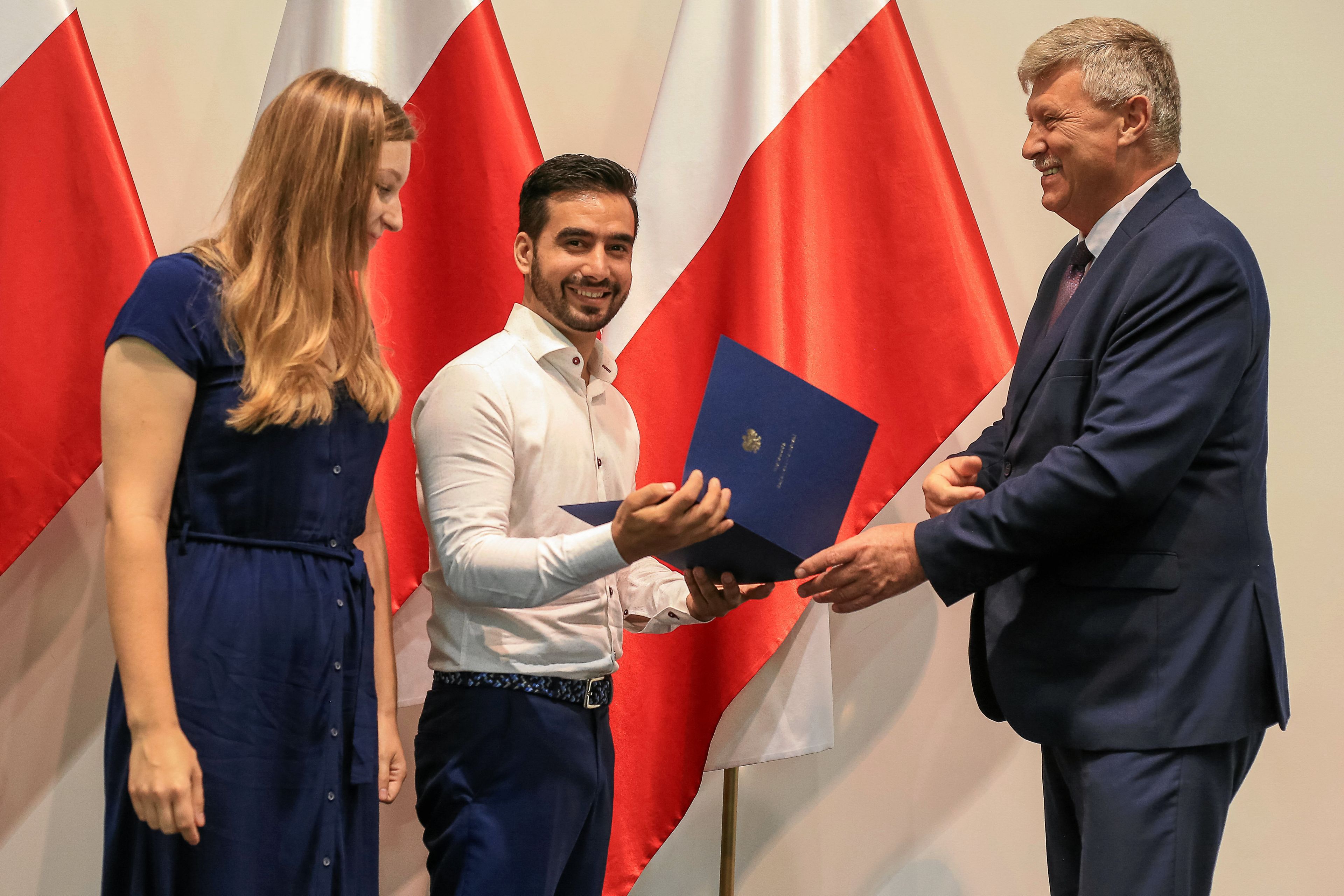 Na zdjęciu jest trzy osoby, po lewej - kobieta, po prawej dwóch mężczyzn. W tle widać flagi biało-czerwone