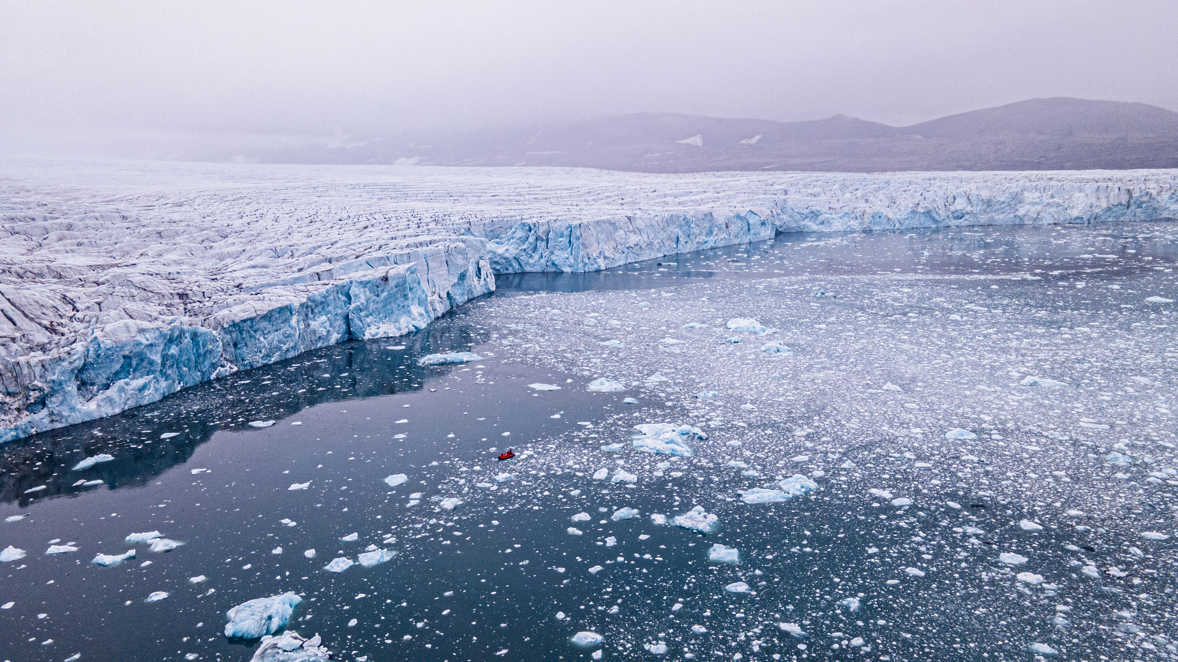 Zdjęcie z góry, widok na ocean i ścianę lodowca, na środku mała, czerwona łódka.