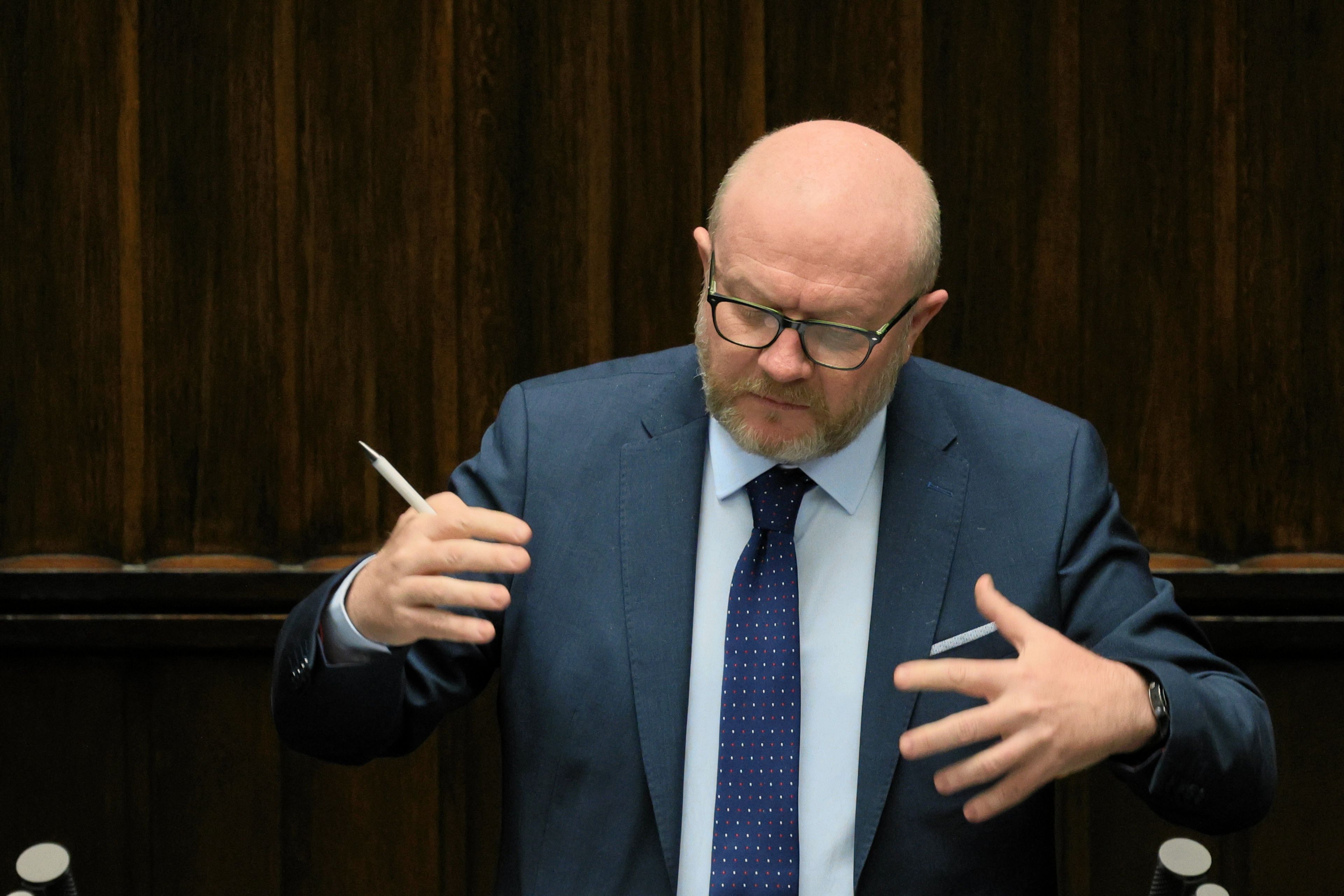 Łysy mężczyzna w garniturze przemawia i gestykuluje.