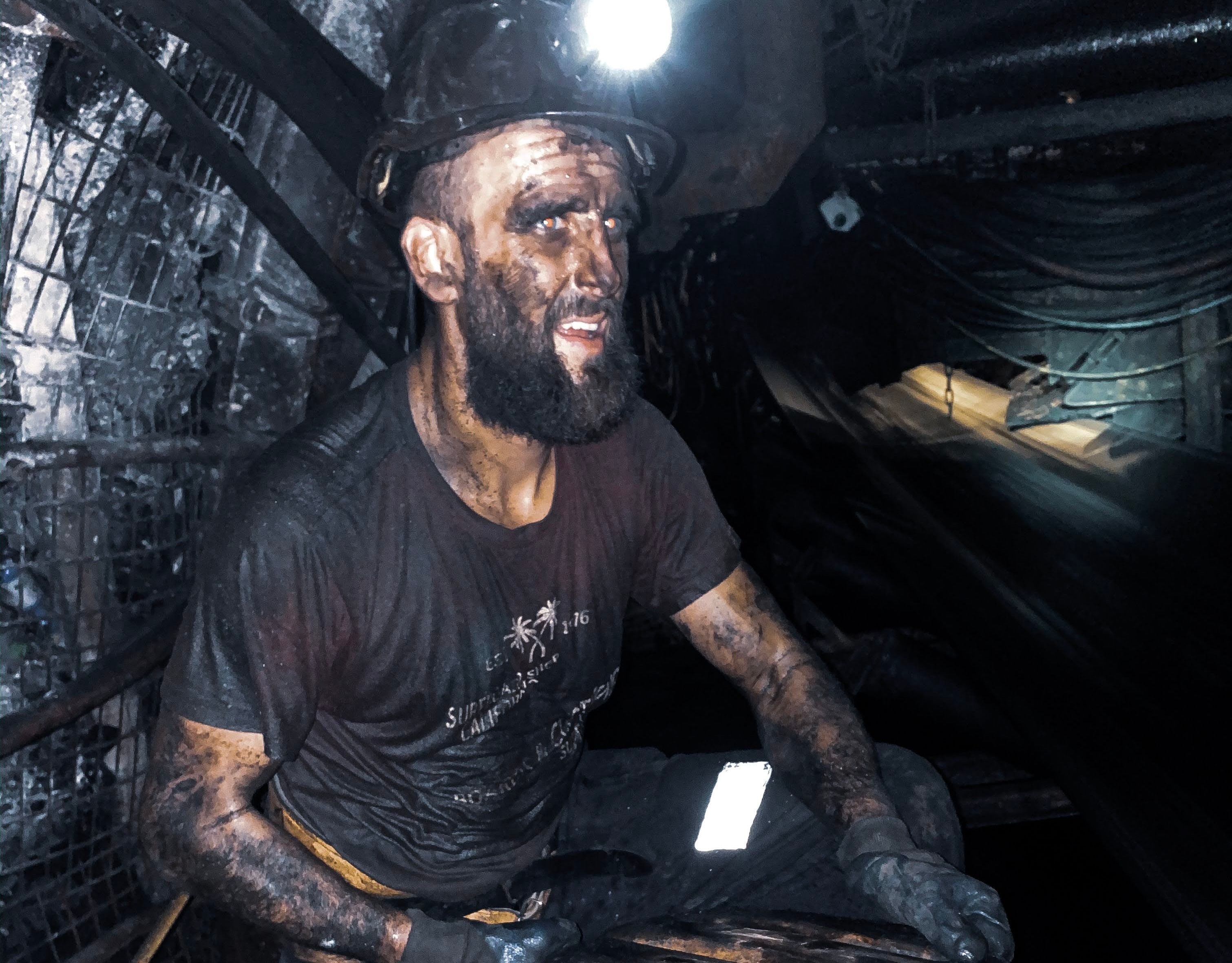 Na zdjęciu młody górnik w podziemiach kopalni. Jest brudny od pyłu węglowego, na głowie ma hełm ze świecącą się czołówką.