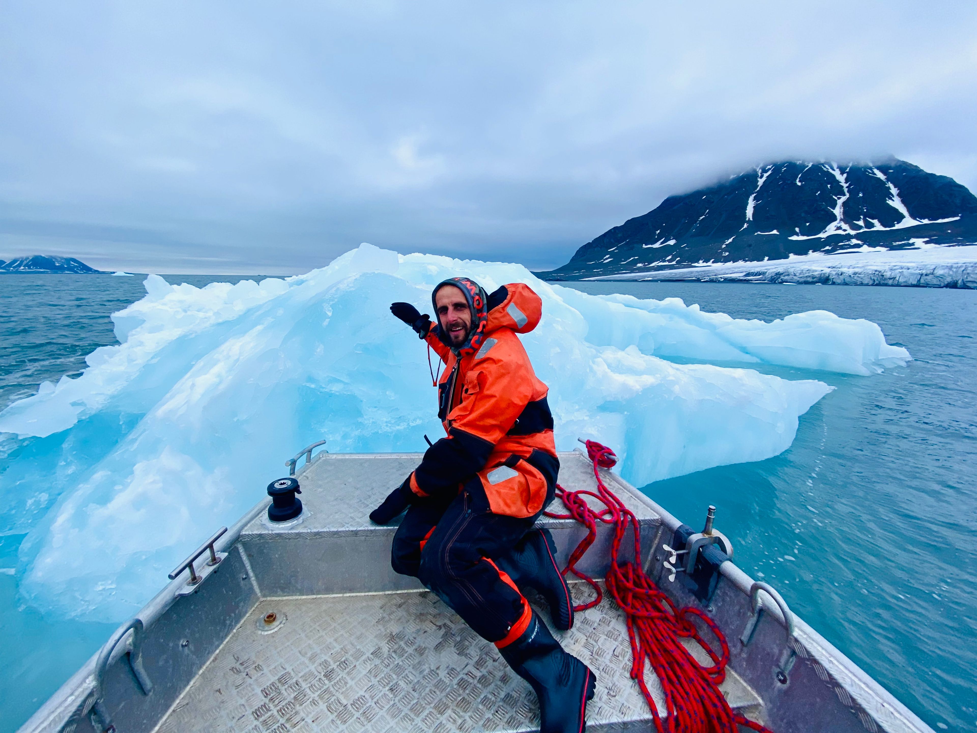 Młody człowiek w pomarańczowym kombinezonie siedzi na łodzi i pokazuje na fragment lodowca.