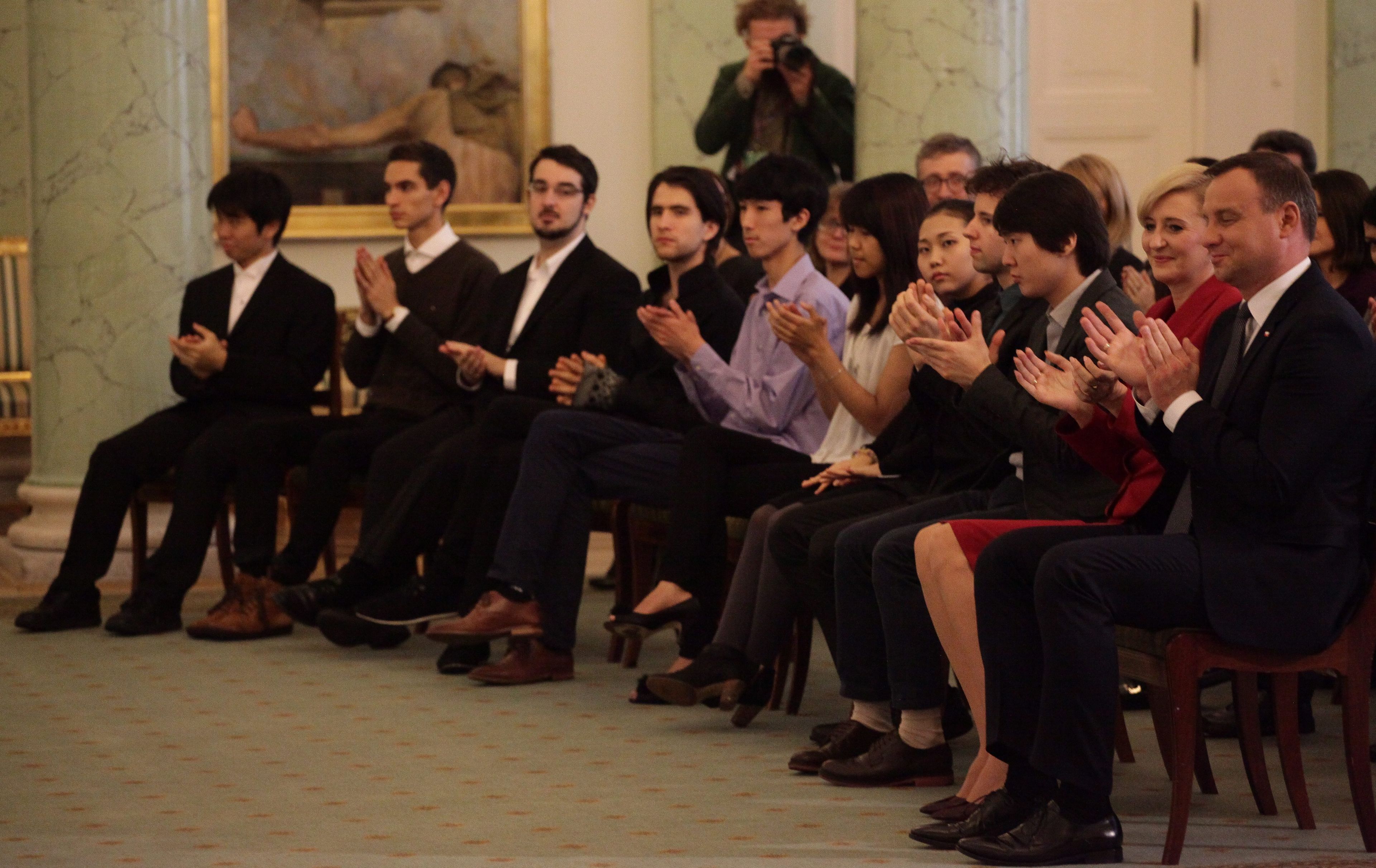 Mlodzi ludzie siedzą w rzędzie. Pośrodku - mężczyzna w szarej koszuli