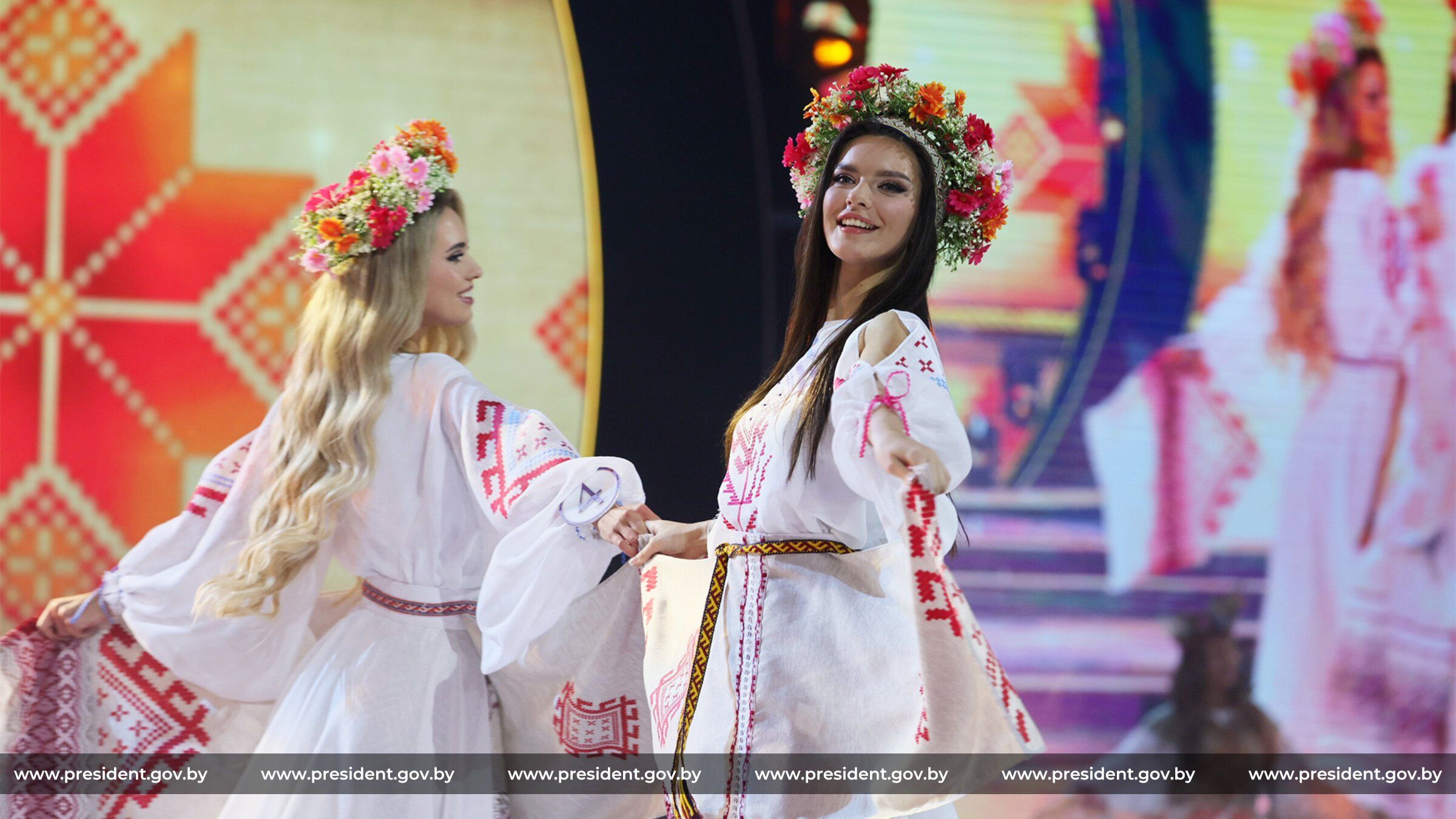Młodziutkie blondyka i brunetka na scenie w wieńcach na głowach i białych ludowych sukienkach z czerwonym haftem.